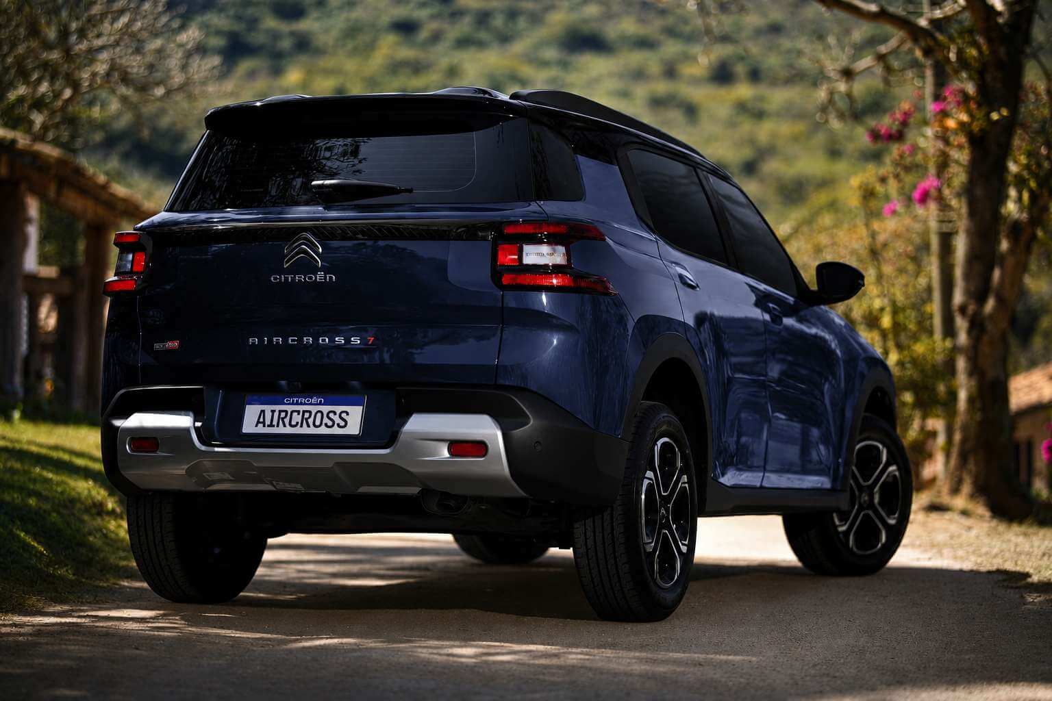 Citroën Aircross 2026 na cor azul-escuro fotografado em tres quartos traseiro, parado em uma estrada de terra de zona rural. A tampa traseira mostra o logotipo de chevrons da Citroën, a palavra CITROËN e a inscricao AIRCROSS 7, com a placa escrita apenas AIRCROSS. Ao redor ha grama, uma cerca de madeira, uma arvore com flores rosa e morros verdes ao fundo, sob luz natural de fim de tarde.