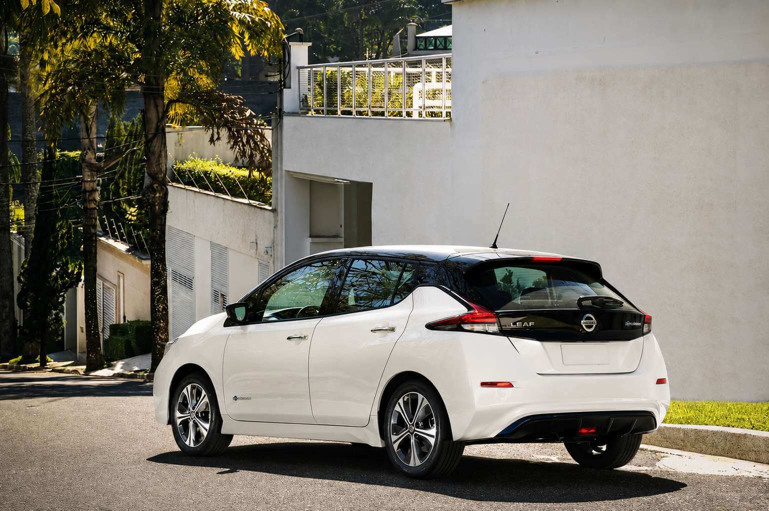 Nissan LEAF branco da geração ZE1 fotografado de três quartos traseiros, parado na beira de uma rua residencial arborizada com palmeiras e muros brancos ao fundo, sob luz forte de fim de tarde. A traseira mostra as lanternas em formato de bumerangue, o emblema da Nissan e o letreiro LEAF na tampa do porta-malas, além das rodas de liga de duas cores; a placa está em branco.