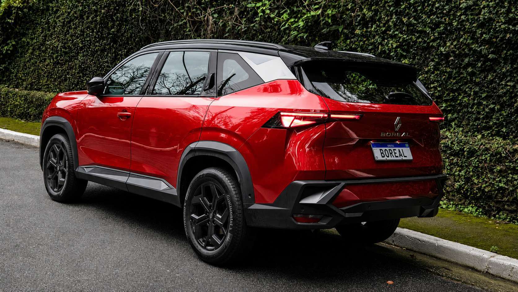 Renault Boreal 2027 na versao Techno, vermelho com teto preto, fotografado de tres quartos traseiro a esquerda, estacionado junto a uma guia de rua em dia nublado e com luz suave. Aparecem a lanterna traseira em faixa horizontal de LED, o losango da Renault e o letreiro BOREAL na tampa do porta-malas, a placa escrita BOREAL, os para-lamas e a parte inferior em plastico preto, as rodas de liga escuras e, ao fundo, uma cerca viva densa de folhagem verde.