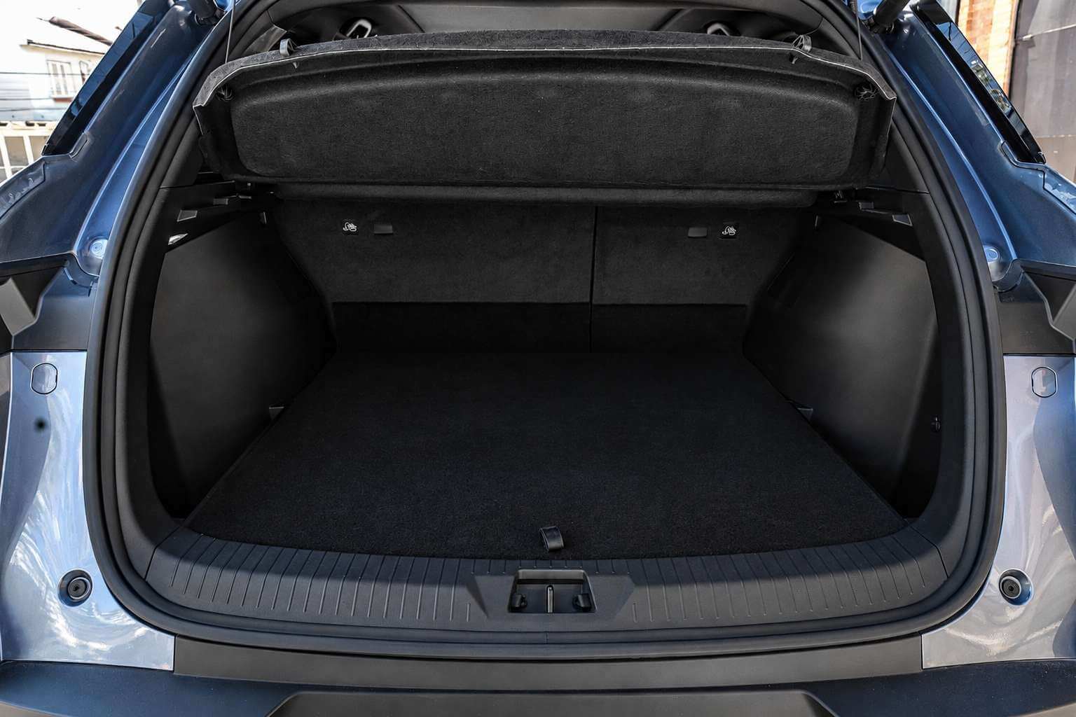 Porta-malas aberto do BYD Song Pro GS 2027, um SUV azul-acinzentado metálico, visto de frente pela tampa traseira levantada. O compartimento é forrado em carpete preto, com assoalho plano e reto, tampa de bagagem rígida presa por cordões logo abaixo do vidro traseiro e encostos traseiros bipartidos ao fundo. Nas laterais há ganchos de amarração e pequenos ícones indicativos; a soleira tem proteção plástica ranhurada. Luz natural do dia entra pelos lados.