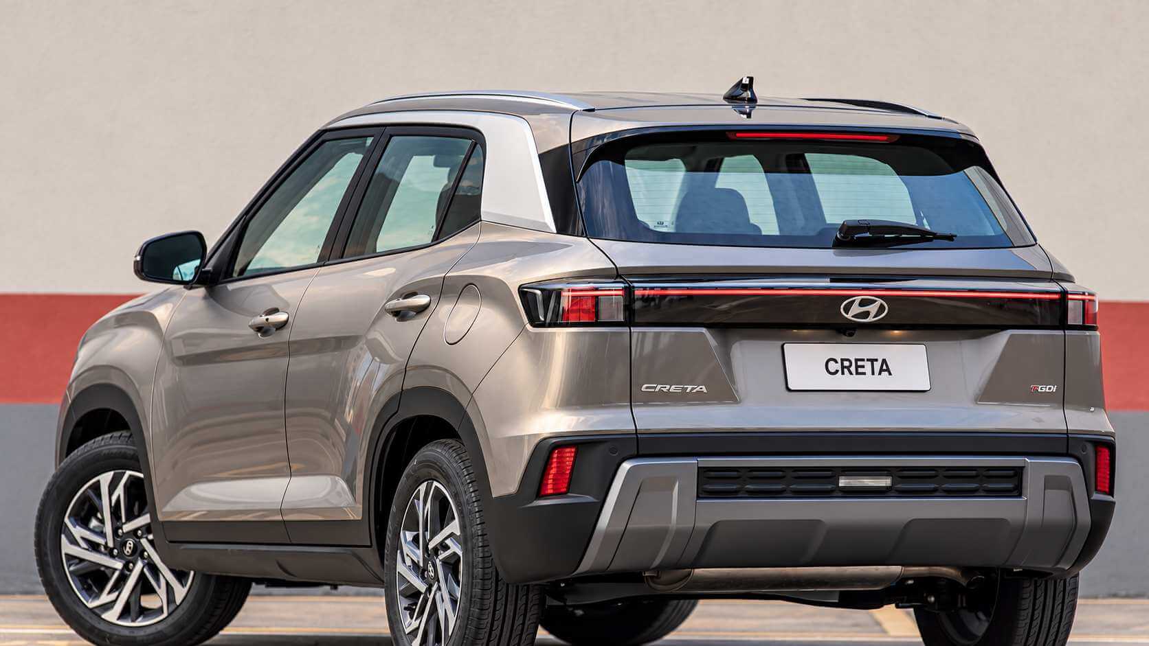 Hyundai Creta cinza-bronze visto de trás em três quartos, parado em um pátio de piso claro com uma faixa vermelha na parede ao fundo. Aparecem a lanterna traseira em faixa contínua, o emblema da Hyundai e o nome CRETA na tampa do porta-malas e a placa traseira com o nome CRETA.