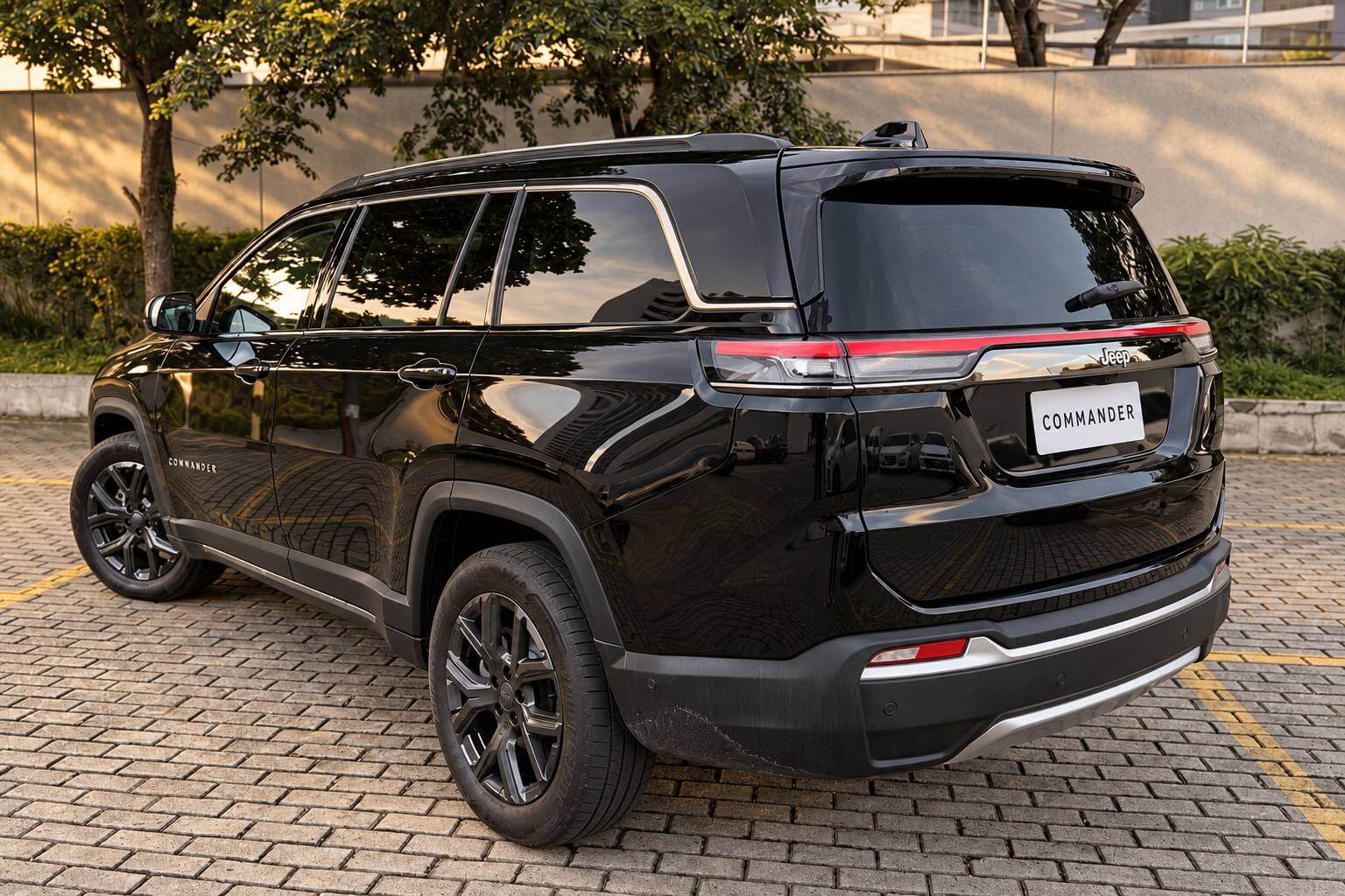 Jeep Commander Longitude preto visto de traseira em três quartos, estacionado sobre piso de bloquete em um pátio arborizado no fim da tarde, com a lanterna traseira em faixa vermelha contínua atravessando a tampa do porta-malas, emblema Jeep centralizado, rodas de liga escuras e placa em branco com a inscrição COMMANDER