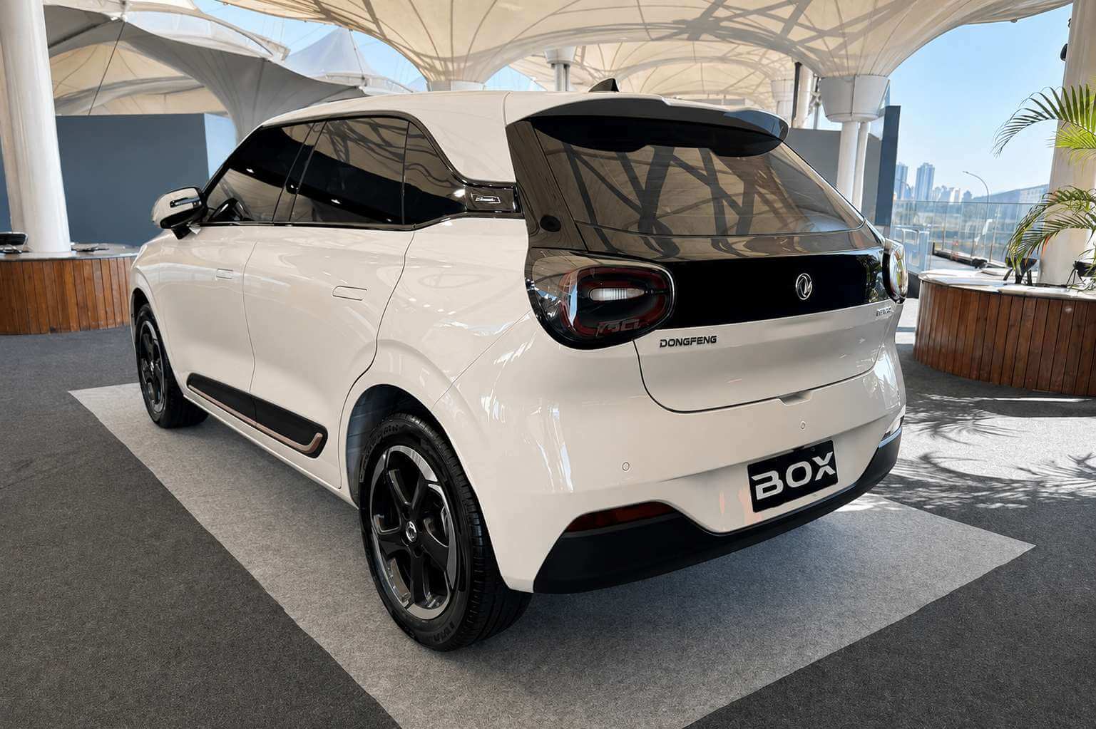 DFM Box Urban 2026, hatch compacto elétrico branco, visto de três quartos traseiro pelo lado esquerdo, em exposição sob as tendas brancas do Festival Interlagos: teto flutuante com antena tipo barbatana, aerofólio e vidro traseiro escurecidos, lanternas de LED unidas por uma faixa preta que atravessa a tampa do porta-malas, emblema da Dongfeng ao centro e o letreiro DONGFENG logo abaixo, placa traseira escrita apenas BOX, rodas de liga leve escuras de raios duplos e frisos pretos com filete claro na lateral; ao fundo, um balcão de madeira, palmeiras e o céu azul ao lado das colunas do autódromo.