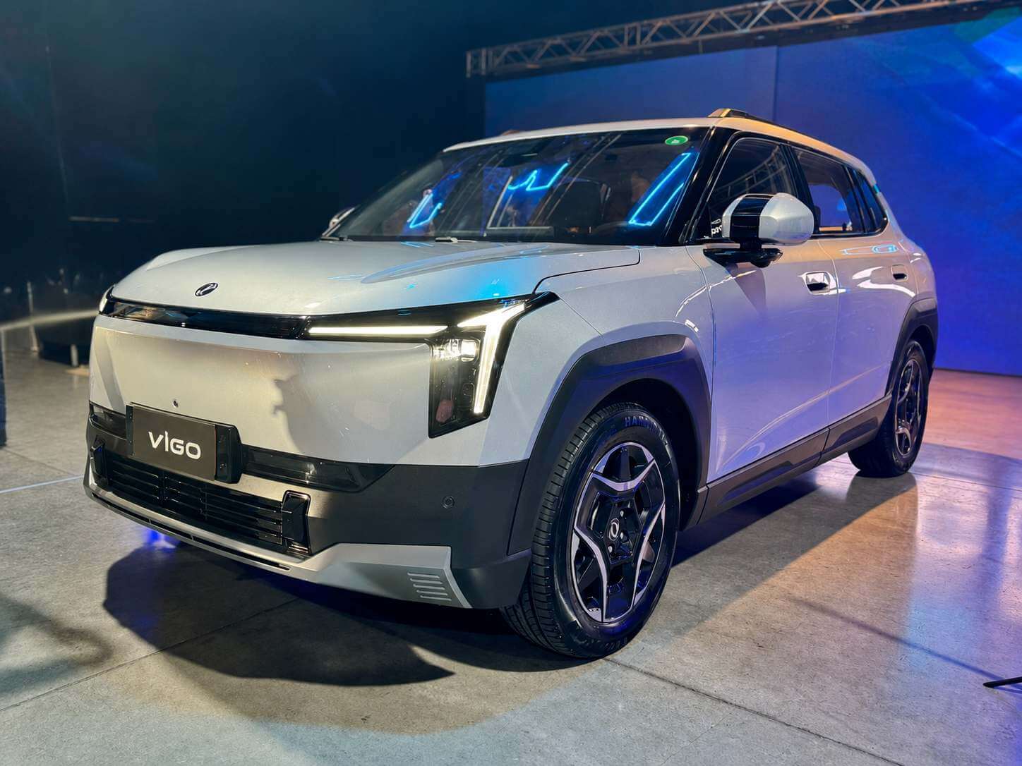 DFM Vigo, o SUV compacto 100% elétrico da marca chinesa, fotografado em três quartos dianteiro. A carroceria é cinza-prata metálico, com para-lamas e saias inferiores em plástico escuro, teto contrastante com friso dourado, rodas de liga leve de raios duplos escuros com detalhes prateados e pneus de perfil alto. Na frente, faróis finos de LED ligados por uma faixa luminosa horizontal, capô alto e liso com o emblema circular da Dongfeng ao centro e uma placa branca em que se lê apenas VIGO. O carro está parado sobre piso de concreto claro em um evento de lançamento fechado, com estrutura metálica de treliça no teto e iluminação azulada ao fundo.