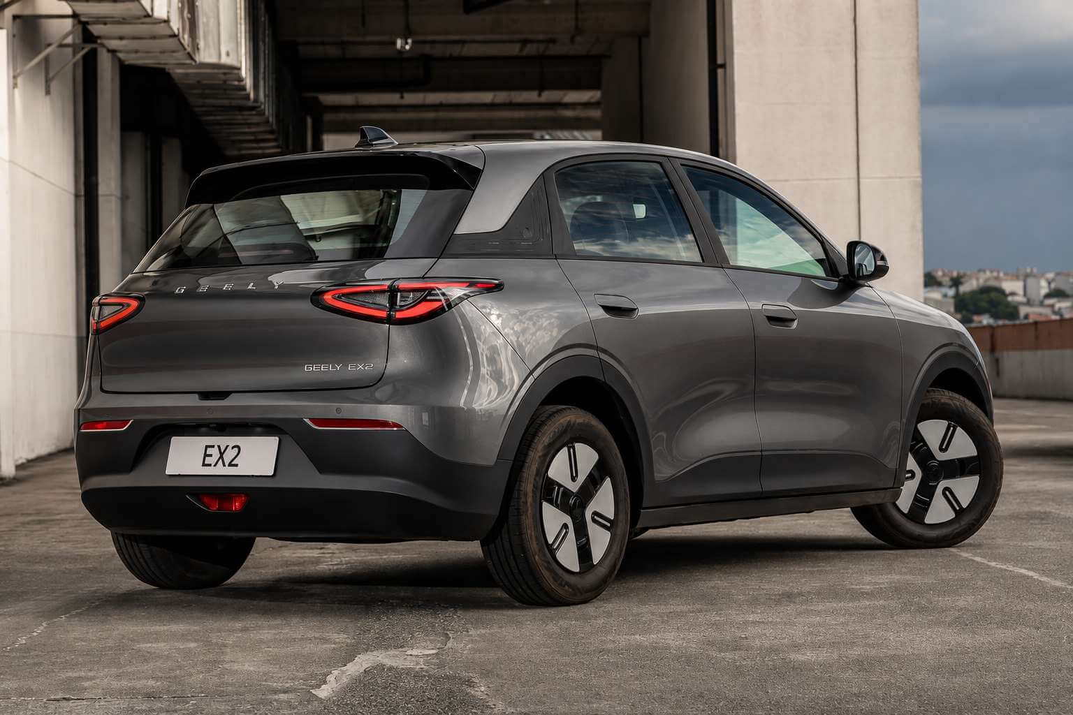 Geely EX2 cinza fotografado de traseira em tres quartos, no piso superior de um estacionamento aberto de concreto. O SUV eletrico compacto aparece com o teto preto em contraste, lanternas traseiras ligadas em formato de C vermelho ligadas por uma faixa escura, o nome GEELY EX2 na tampa do porta-malas e placa branca com a inscricao EX2. As rodas sao aerodinamicas em duas cores, pretas com aletas brancas. Ao fundo, colunas de concreto e o ceu nublado sobre a cidade.