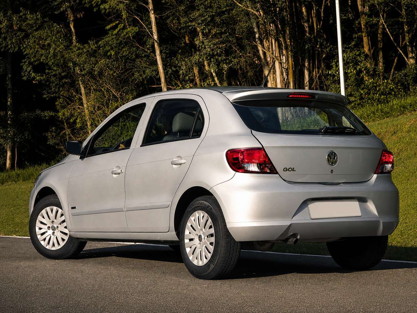 Volkswagen Gol G5 1.0 prata, hatch de entrada, visto de traseira em três quartos numa estrada de asfalto com mata verde ao fundo sob luz de fim de tarde; rodas de aço com calotas, emblema VW e letreiro GOL na tampa traseira