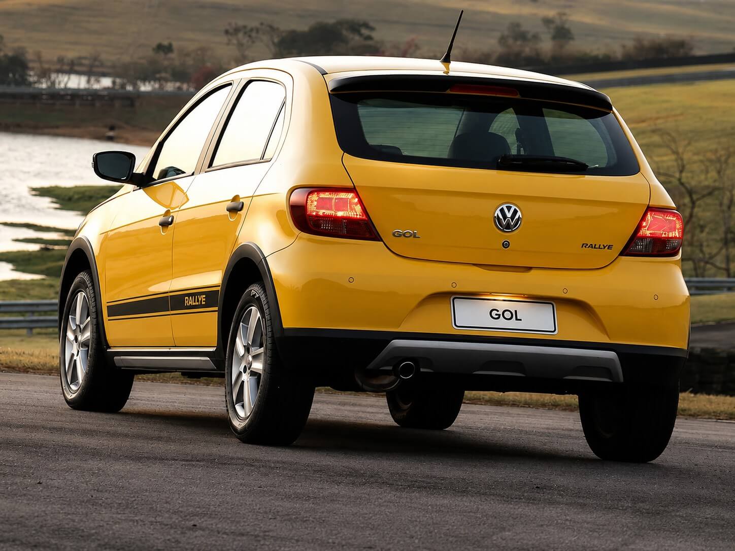 Volkswagen Gol Rallye 1.6 amarelo visto de traseira em três-quartos, numa estrada de asfalto à beira de um lago no campo ao entardecer; molduras pretas nos para-lamas, suspensão elevada, rodas de liga aro 15 e o adesivo RALLYE na lateral