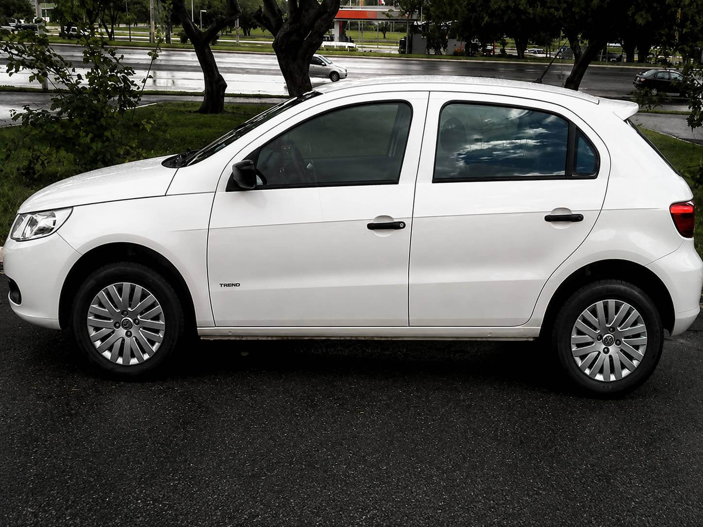 Volkswagen Gol G5 Trend 1.0 branco, hatch de entrada, visto de dianteira em três quartos parado numa rua urbana com parede azul e branca ao fundo; rodas aro 14 com calotas, maçanetas na cor da carroceria, emblema VW na grade e placa escrita GOL