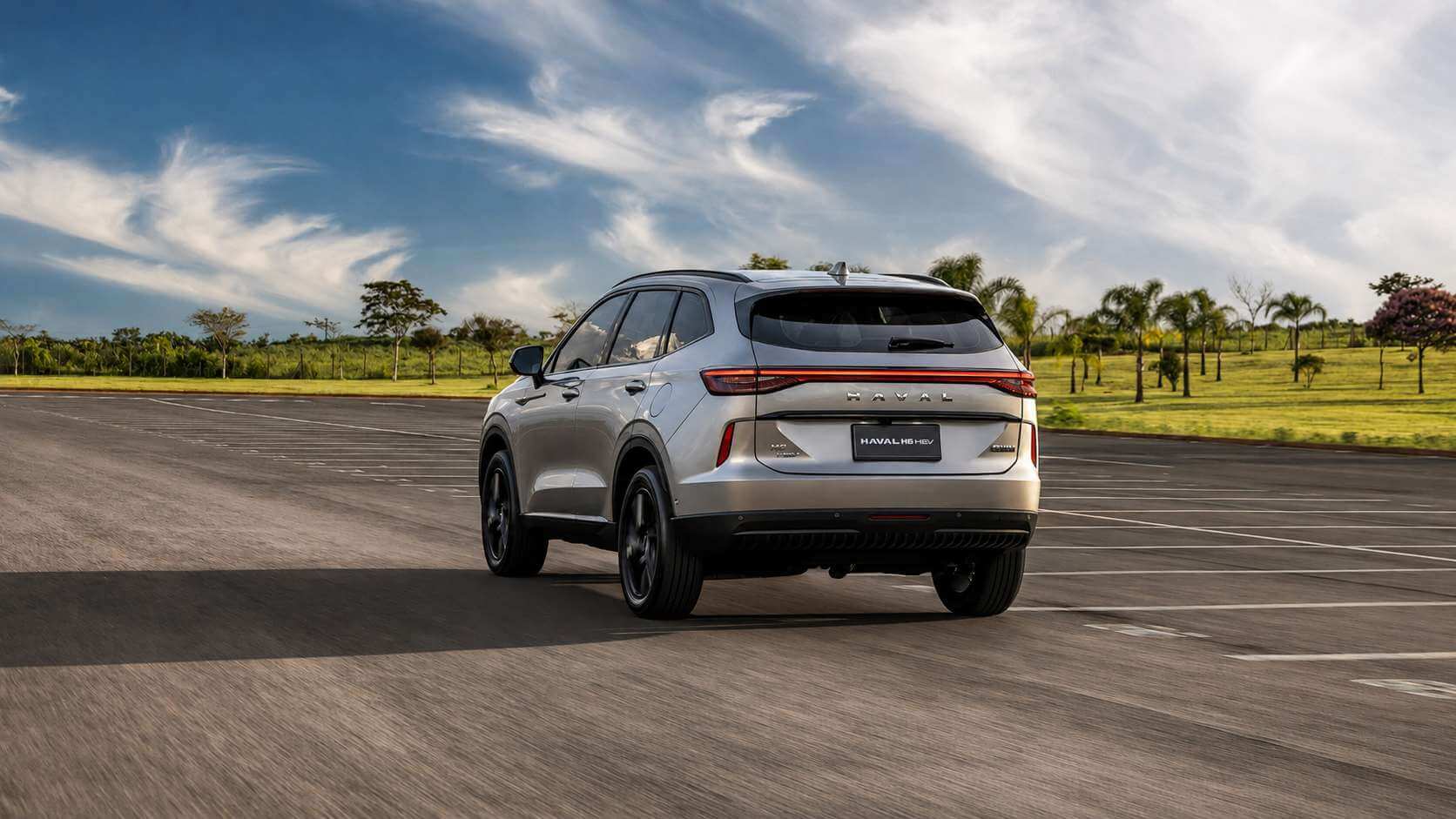 GWM Haval H6 HEV prateado visto de trás em três quartos, rodando por uma pista de testes com gramado e árvores ao fundo no fim da tarde. A faixa de lanternas que atravessa a tampa do porta-malas, o letreiro HAVAL no centro e as rodas escurecidas aparecem em destaque.