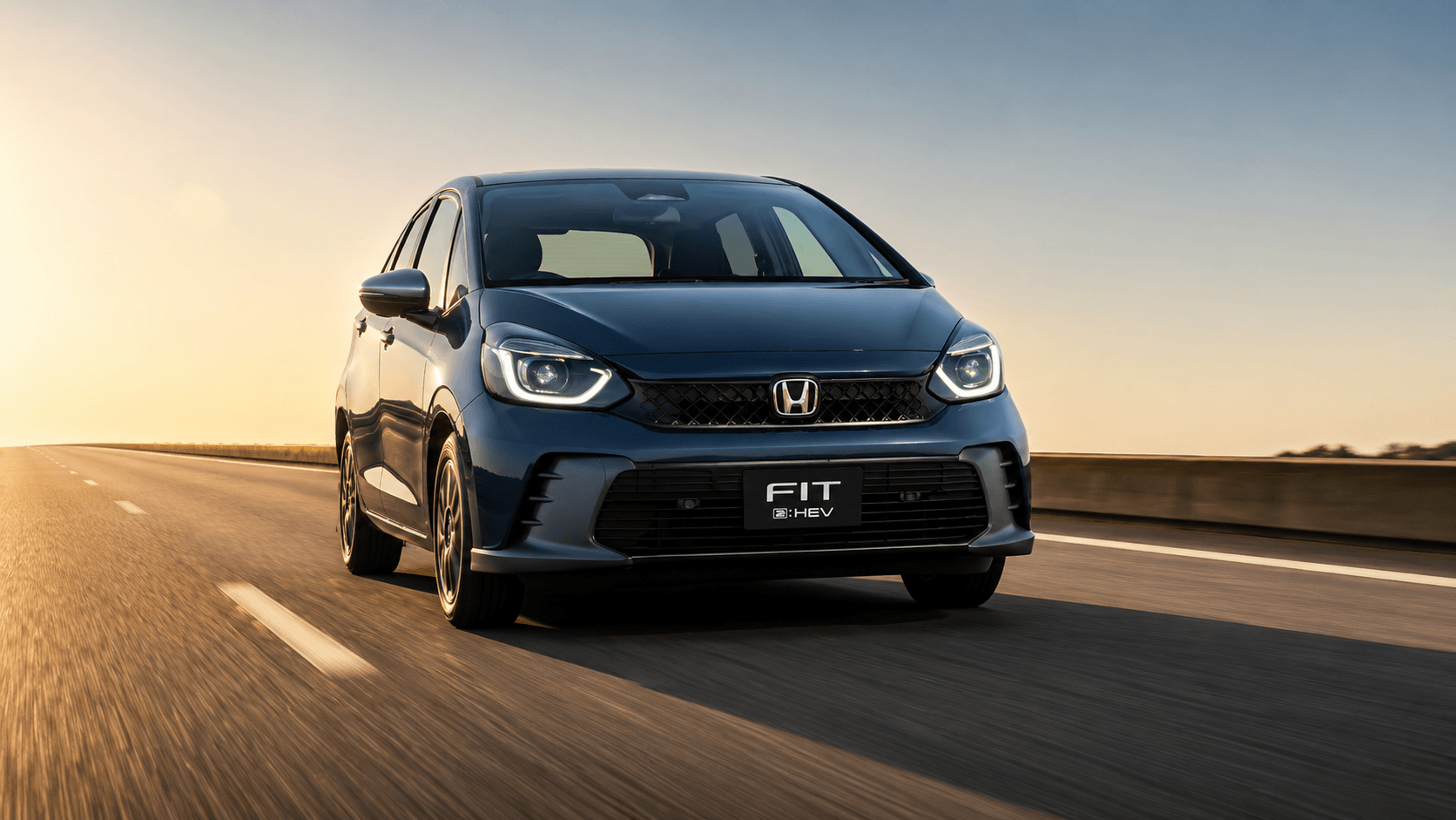 Honda Fit e:HEV azul-petróleo visto de frente em três quartos, rodando numa rodovia ao pôr do sol com o asfalto desfocado pela velocidade; faróis de LED acesos e grade preta com o logo da Honda.