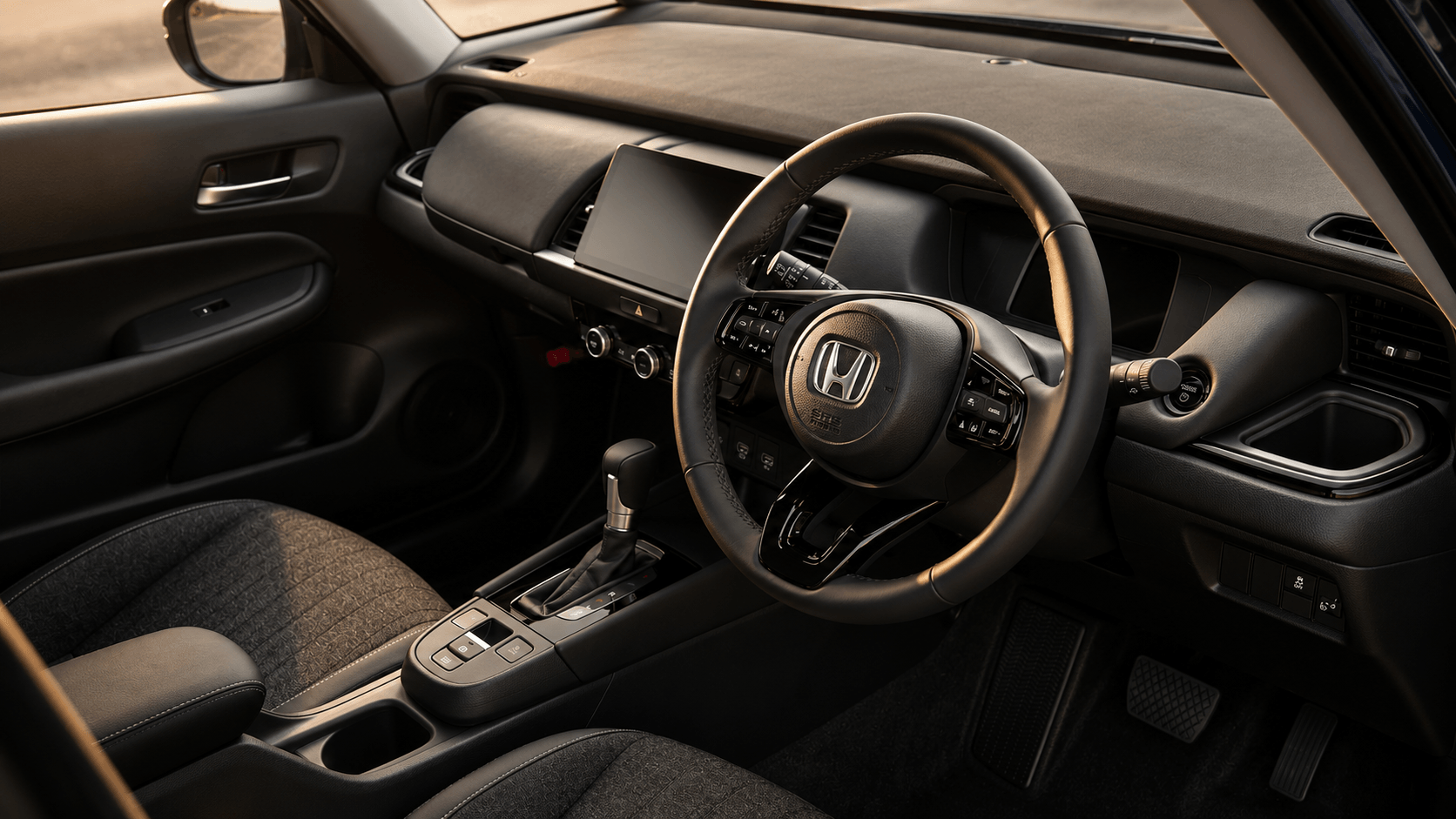 Interior preto do Fit com o volante à direita, no padrão japonês; central multimídia vertical no alto do painel, alavanca do câmbio automático no console e bancos revestidos em tecido texturizado, iluminados por uma luz quente.