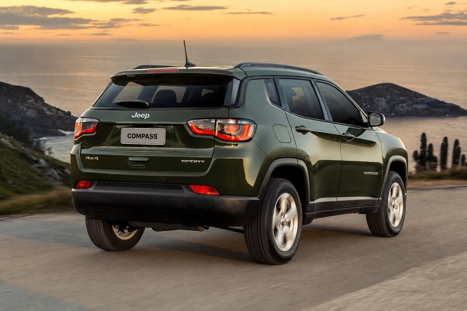 Jeep Compass verde-escuro visto de tras em tres quartos, rodando por uma estrada de asfalto no alto de uma encosta a beira-mar ao entardecer, com o ceu alaranjado, o mar e um morro rochoso ao fundo e cactos na beira da pista. Aparecem o emblema Jeep na tampa traseira, os badges 4x4 e SPORT, o letreiro COMPASS na lateral, as lanternas acesas e as rodas de liga prateadas.