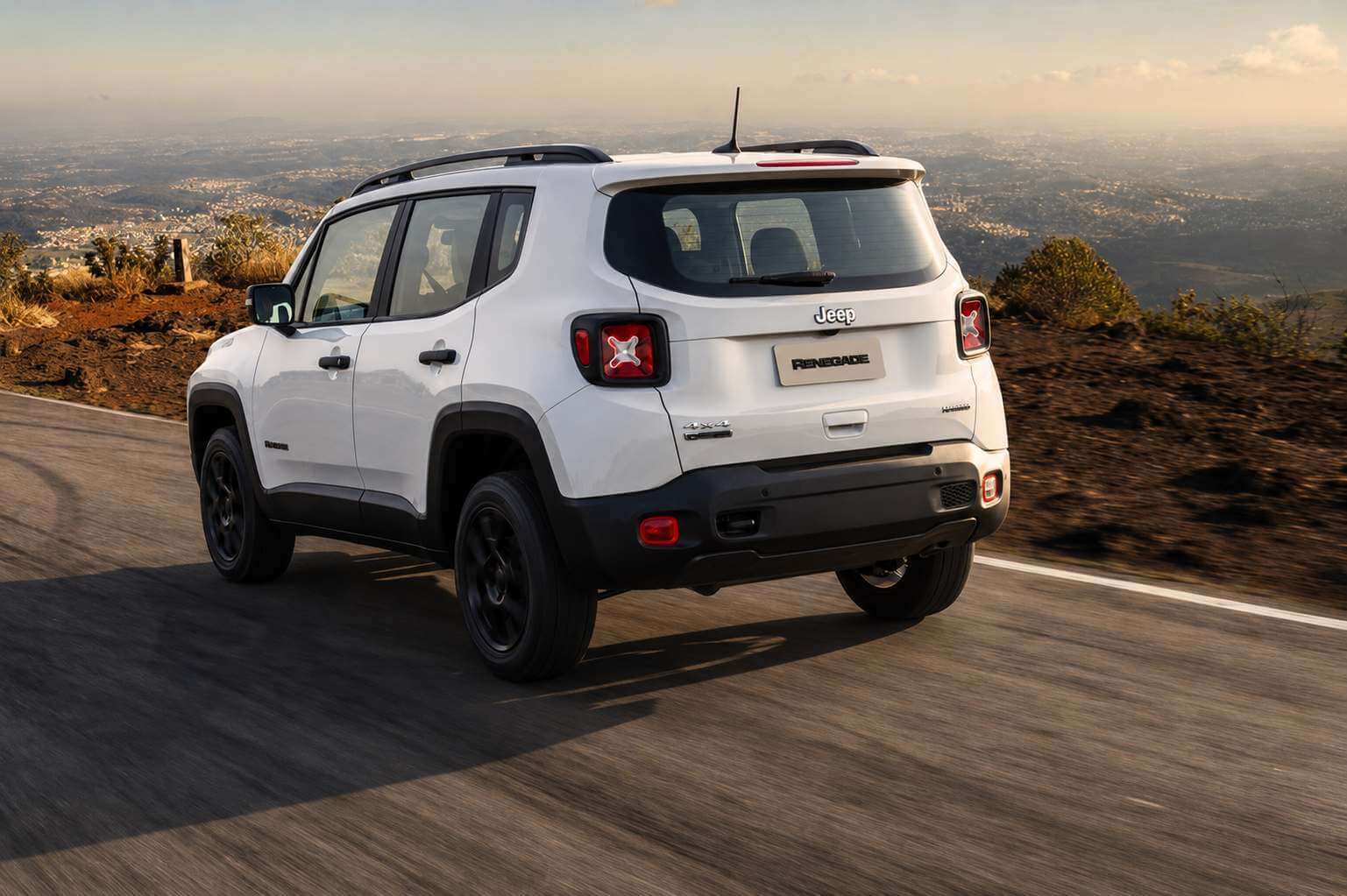 Jeep Renegade branco visto de trás em três quartos, rodando por uma estrada de montanha no fim da tarde; as lanternas em formato de X, o emblema Jeep na tampa traseira e a placa escrita RENEGADE aparecem em destaque, com o vale iluminado pela luz dourada ao fundo.