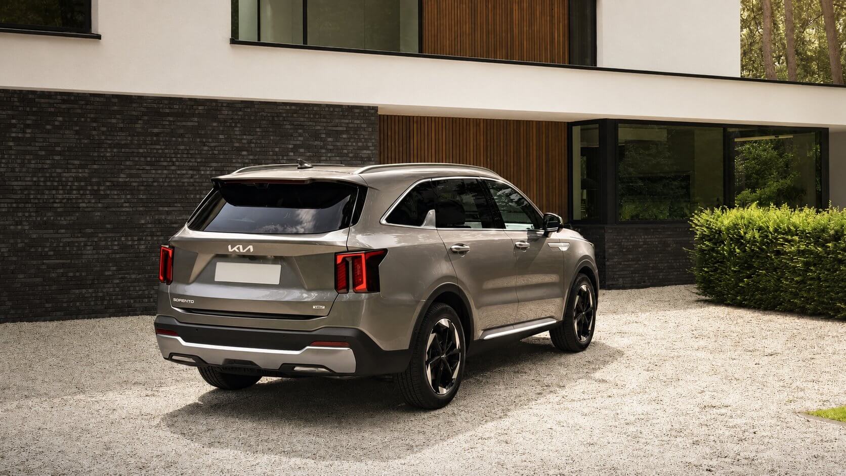 Kia Sorento EX 2026 cinza visto de tras em angulo tres-quartos, parado na entrada de cascalho de uma casa moderna, com as lanternas verticais de LED em assinatura e rodas de liga escuras.