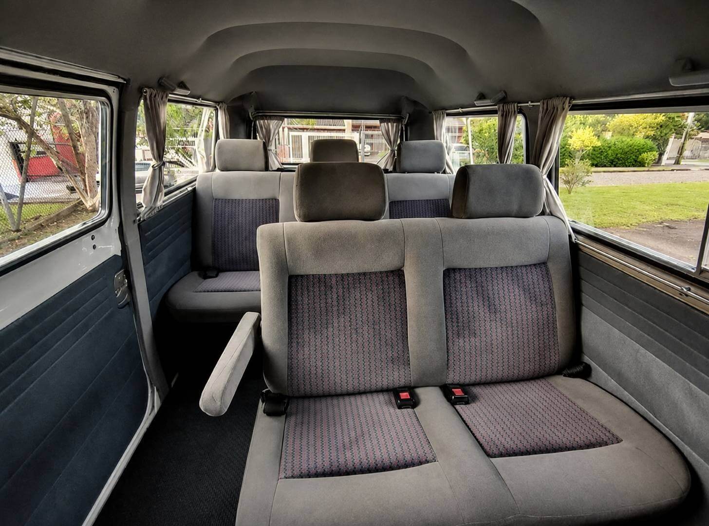 Interior de uma Volkswagen Kombi vista de dentro, a partir do banco traseiro, em direcao a frente do veiculo. Os bancos sao estofados em tecido cinza claro com painel central xadrez em tons de cinza escuro e vinho; na frente aparece um banco corrido com dois encostos de cabeca e, mais a frente, os bancos dianteiros, tambem com encostos de cabeca. A esquerda ha um banco lateral menor junto a porta e um apoio de braco estofado dobrado para baixo. O forro do teto e cinza claro, o assoalho e coberto por carpete escuro e os painies das portas combinam plastico claro com estofamento azul escuro. As janelas laterais tem cortinas bege presas nas laterais e, atraves dos vidros, veem-se arvores, grama verde e uma rua residencial em dia nublado. Dois cintos de seguranca com fivelas vermelhas aparecem no banco traseiro.