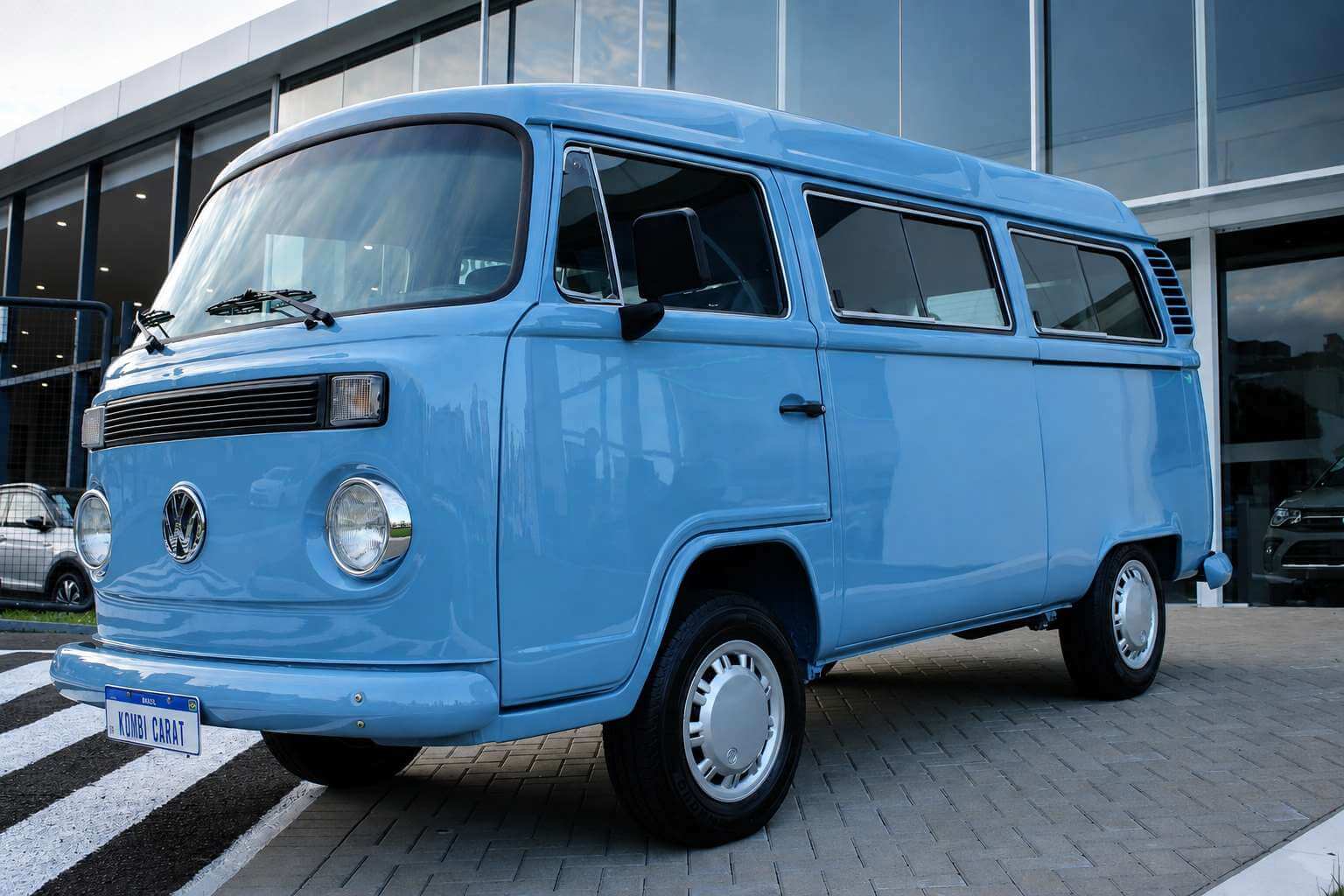 Volkswagen Kombi Carat 1999 na cor azul claro, restaurada, fotografada em tres quartos dianteiro a partir de um angulo baixo no patio de uma concessionaria. A frente chata da ultima geracao a ar aparece inteira: o emblema VW cromado no centro do bico, os dois farois redondos com aros cromados, a grade preta de ventilacao acima do para-choque e a placa branca com os dizeres KOMBI CARAT. A lateral direita mostra a porta corredica, as janelas com molduras cromadas e as calotas claras sobre pneus de faixa preta. Ao fundo, a fachada de vidro da loja reflete o ceu do fim de tarde.