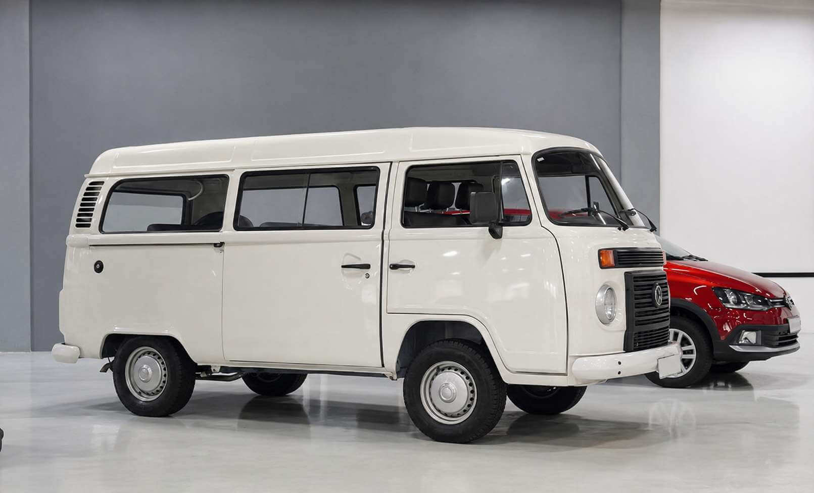Volkswagen Kombi Standard 1.4 Flex branca, geração final da linha, fotografada em três quartos dianteiro num showroom de piso claro e paredes cinza; teto elevado, frente plana com faróis quadrados, emblema VW cromado na grade preta e rodas de aço com calotas prateadas. Ao fundo, à direita, aparece um Volkswagen Gol vermelho.