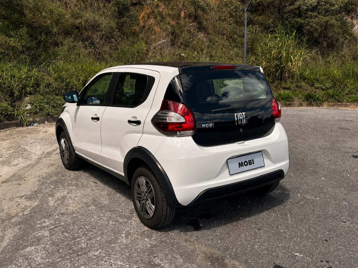Fiat Mobi Like branco visto de tres quartos traseiro, parado no acostamento de uma estrada de asfalto com barranco de vegetacao verde ao fundo, sob luz do dia; a tampa traseira mostra o letreiro FIAT e o nome mobi, e a placa traz apenas a palavra MOBI