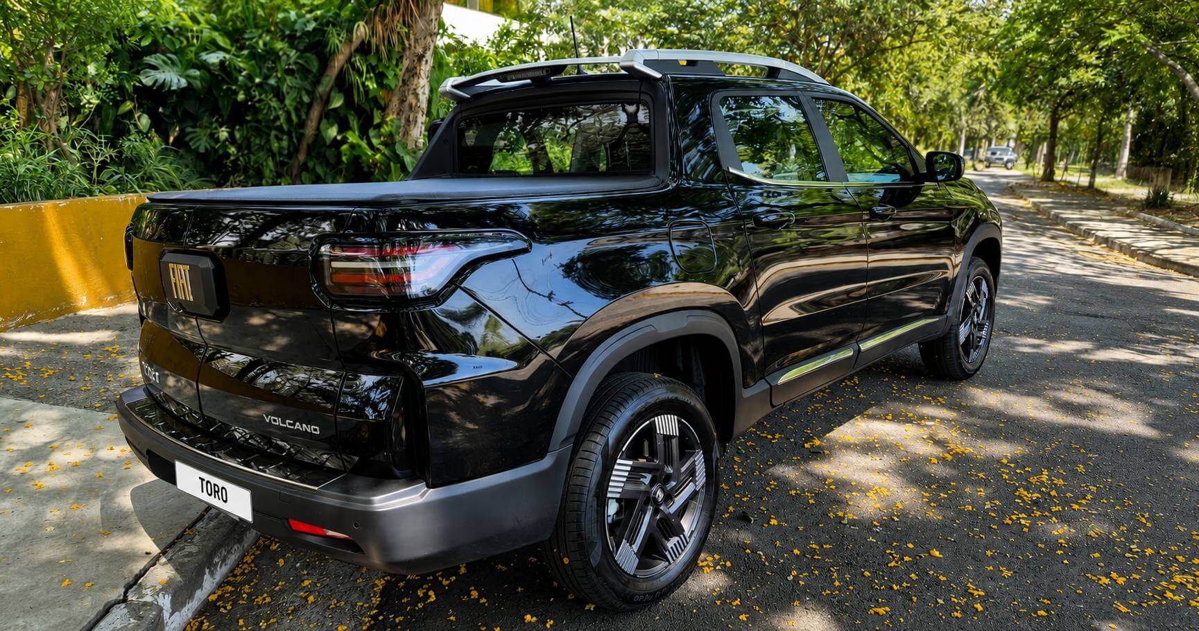 Traseira em tres quartos da Fiat Toro 2026 Volcano preta, estacionada na beira de uma rua arborizada coberta de petalas amarelas de ipe. A pintura escura reflete as copas das arvores; na traseira reta da versao reestilizada aparecem o emblema FIAT em relevo no centro da tampa da cacamba, as lanternas horizontais em LED unidas por uma faixa escura, o letreiro VOLCANO e as rodas de liga escuras de cinco raios.