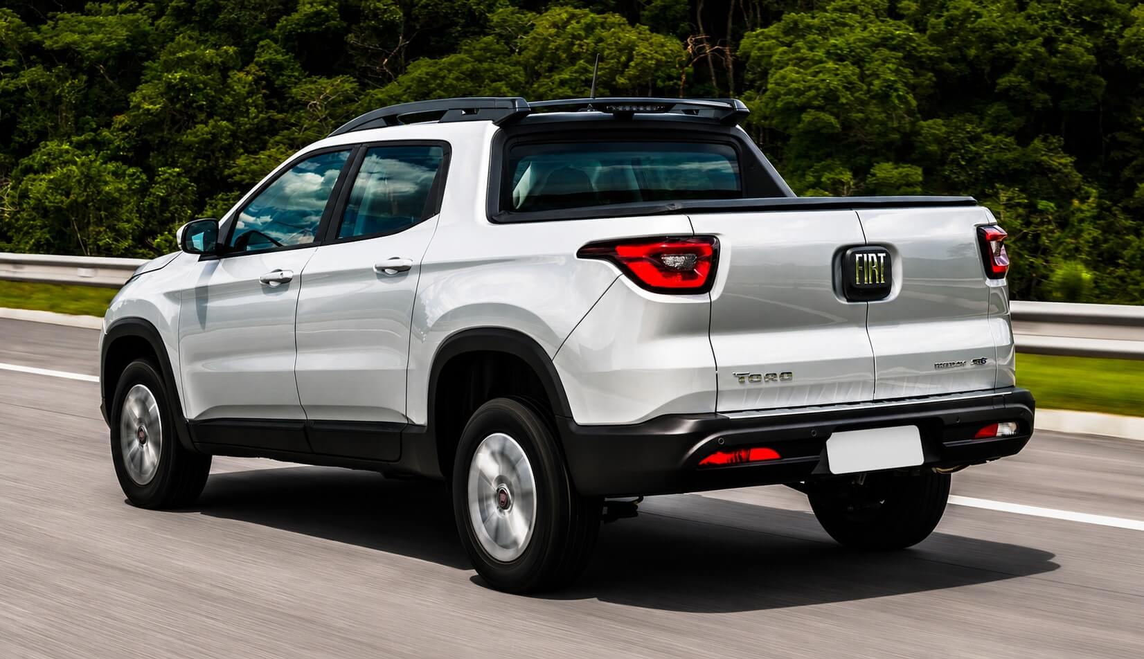 Traseira em tres quartos da Fiat Toro branca da geracao anterior a reestilizacao, a mesma vendida de 2016 a 2021, rodando por uma estrada de pista dupla com a mata verde fechada ao fundo. O carro esta em movimento e o asfalto aparece levemente borrado sob as rodas. Na tampa da cacamba veem-se a letra em relevo TORO do lado esquerdo, o emblema quadrado preto da Fiat no centro e a inscricao menor FREEDOM AT6 do lado direito, que identifica a versao 1.8 flex automatica. As lanternas traseiras sao separadas, em formato retangular arredondado e de cor vermelha, sem a barra de LED que liga as duas na versao reestilizada. A picape tem barras longitudinais no teto, santantonio sobre a cacamba, rodas de liga leve prateadas de cinco raios e a placa esta em branco. Luz de dia, ceu claro refletido nos vidros.