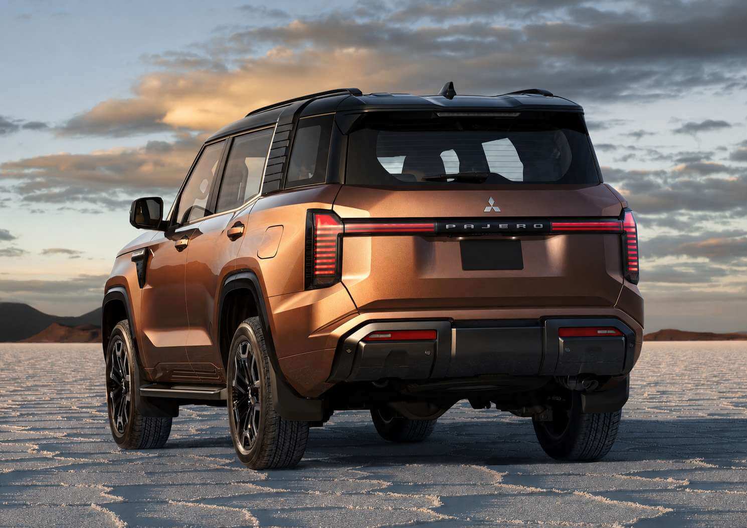 Traseira em tres quartos do novo Mitsubishi Pajero 2027, um SUV grande de carroceria marrom-acobreada metalica com teto preto, parado sobre um deserto de sal claro e rachado. A tampa traseira e limpa, sem estepe pendurado, e traz o emblema dos tres diamantes da Mitsubishi acima de uma faixa de LED vermelha que atravessa toda a largura do carro, com o nome PAJERO em letras espacadas no centro. As rodas sao de liga escura, o para-choque inferior e preto texturizado e ao fundo aparecem montanhas baixas sob um ceu de fim de tarde com nuvens douradas.