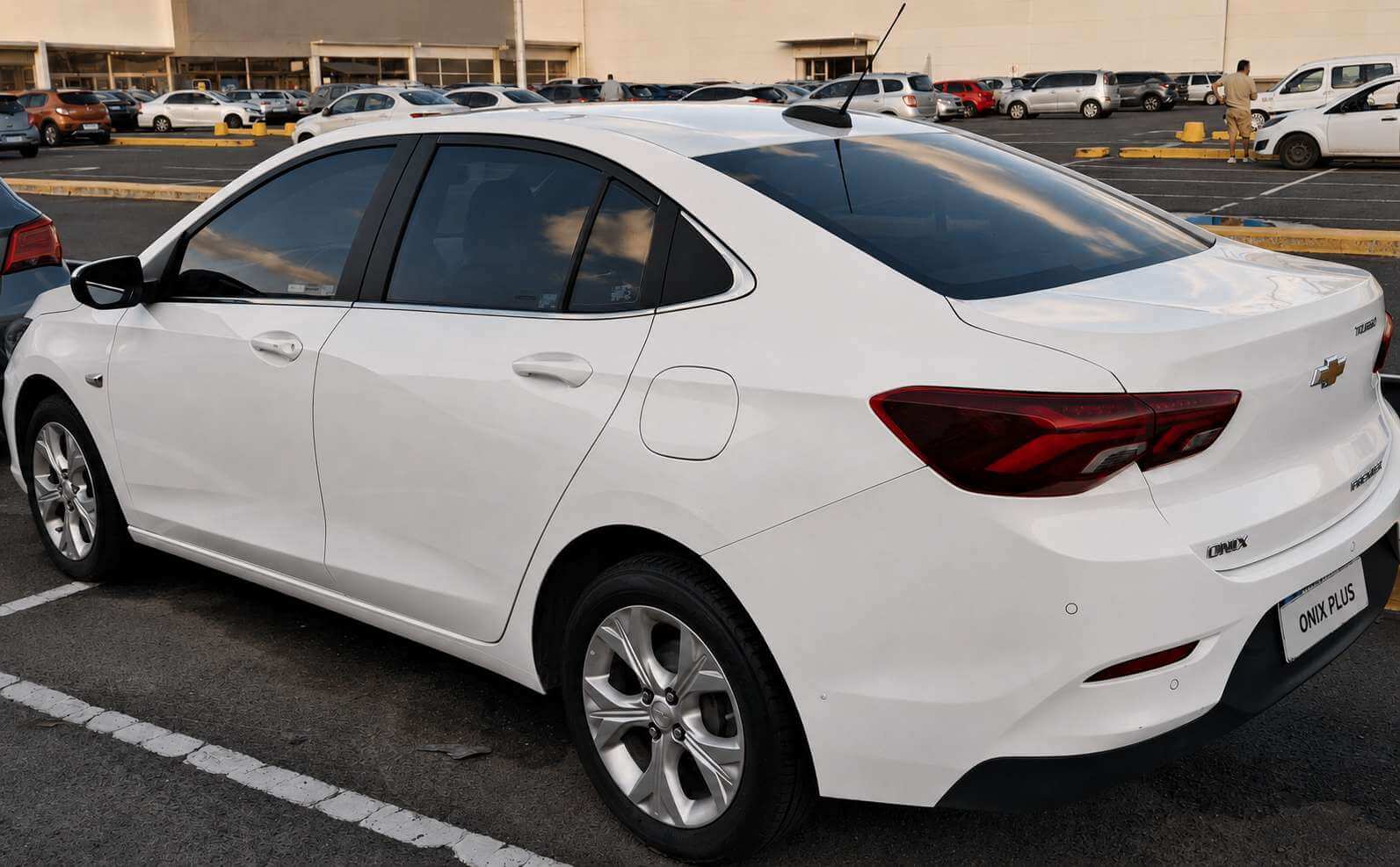 Chevrolet Onix Plus Premier branco, sedã compacto de quatro portas, visto em ângulo três-quartos traseiro, estacionado em uma vaga demarcada de um estacionamento aberto sob a luz natural do fim de tarde. Aparecem a lanterna traseira em LED de recorte alongado, a gravata cromada da Chevrolet no centro da tampa do porta-malas, os emblemas ONIX, PREMIER e TURBO, as rodas de liga leve de cinco raios duplos e os vidros traseiros escurecidos. A placa está em branco, com apenas as palavras ONIX PLUS. Ao fundo, outros carros estacionados e um prédio comercial claro, sem letreiros legíveis.