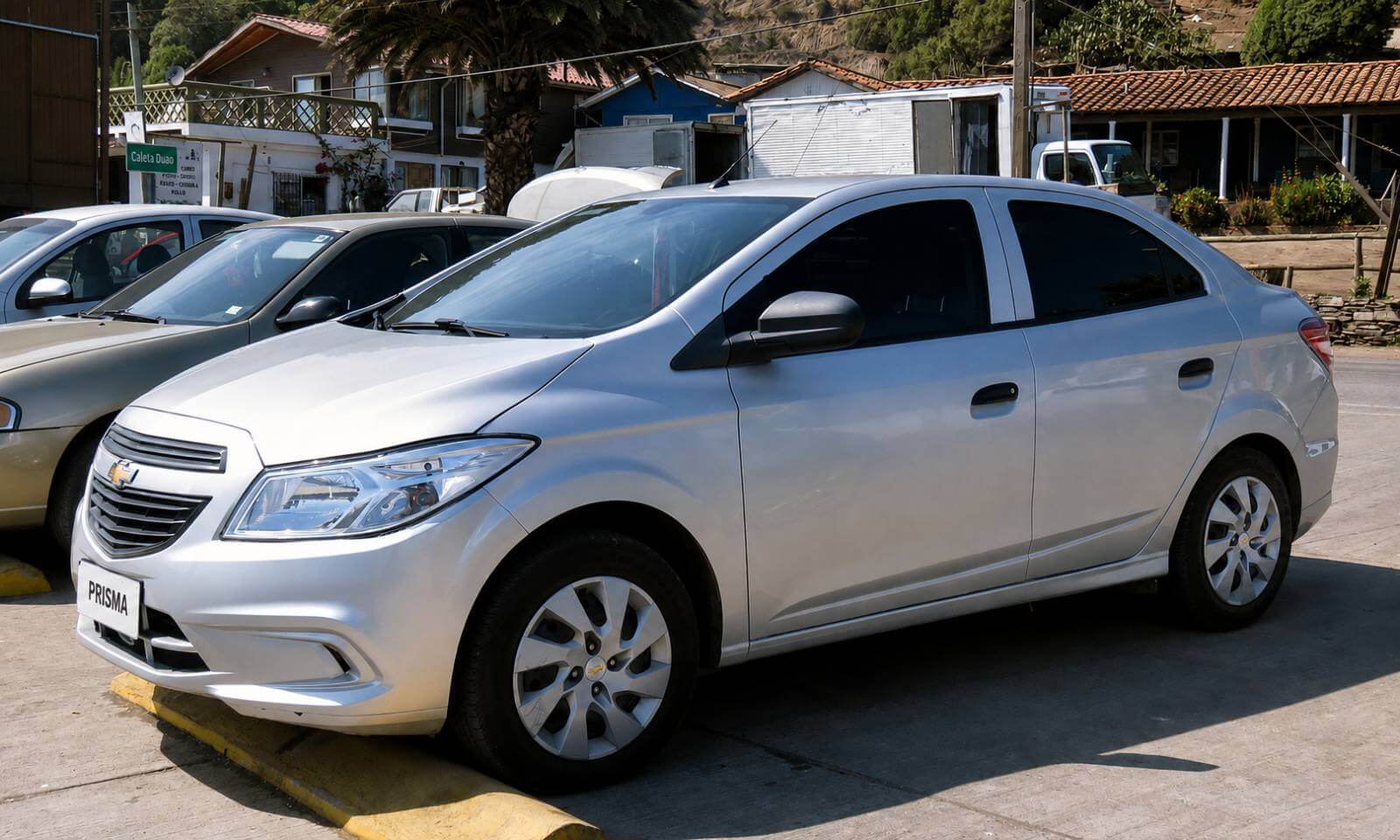 Chevrolet Prisma LT 1.4 2017 prata, visto de tres-quartos dianteiro, estacionado em uma rua de calcada de concreto sob sol forte; o seda compacto exibe a grade preta com o emblema dourado da Chevrolet, farois com mascara escura, calotas de aco prateadas e vidros traseiros escurecidos, com casas simples de telhado de barro e palmeiras ao fundo