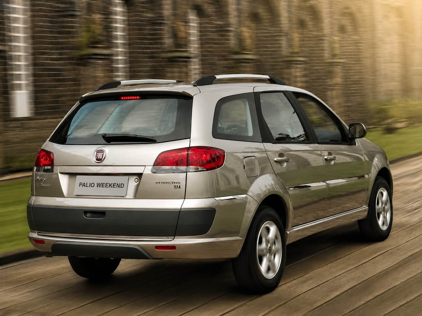 Fiat Palio Weekend Attractive 1.4 2016 na cor prata metálico, vista traseira em três quartos, em movimento sobre um piso de madeira ao lado de um muro de pedra com arcos ao fundo; aparecem as lanternas traseiras vermelhas, o emblema da Fiat e a inscrição Palio Weekend na tampa do porta-malas, a terceira luz de freio, as barras longitudinais no teto e as rodas de liga leve, sob luz quente de fim de tarde.