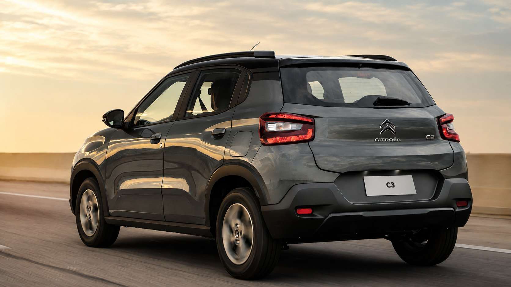 Citroën C3 Feel 1.0 cinza, geração brasileira do hatch, visto de traseira em três quartos enquanto roda por uma estrada de asfalto ao fim da tarde. A luz dourada do sol baixo reflete na lataria; aparecem as lanternas verticais vermelhas, o logo de duplo chevron e o letreiro CITROËN na tampa traseira, o emblema C3 à direita, as barras longitudinais no teto preto e as rodas de liga leve escuras. A placa mostra apenas C3 e o fundo traz o horizonte desfocado pelo movimento.