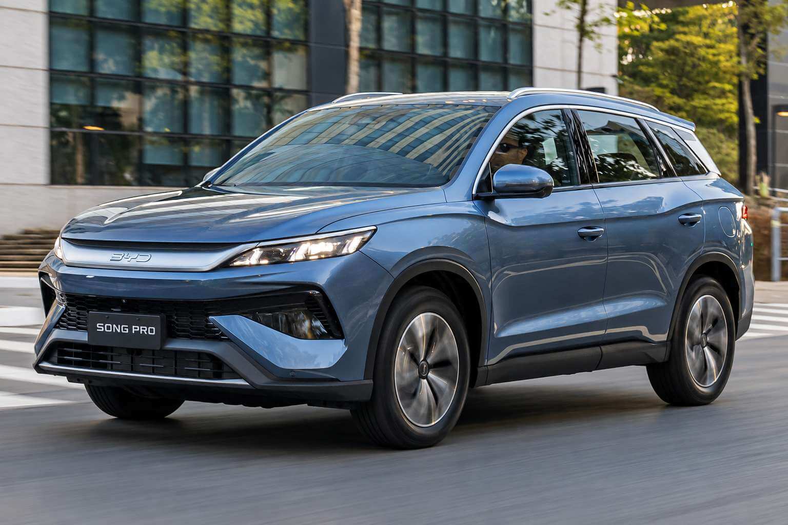 BYD Song Pro GS 2026 na cor azul-acinzentada, vista de tres quartos dianteira, rodando por uma rua urbana arborizada em frente a um predio de fachada envidracada. O SUV hibrido plug-in flex aparece em movimento, com os farois de LED acesos, grade fechada com o logotipo BYD em destaque, rodas de liga leve de dois tons, barras longitudinais no teto e placa dianteira escrita SONG PRO, sob luz natural de fim de tarde.