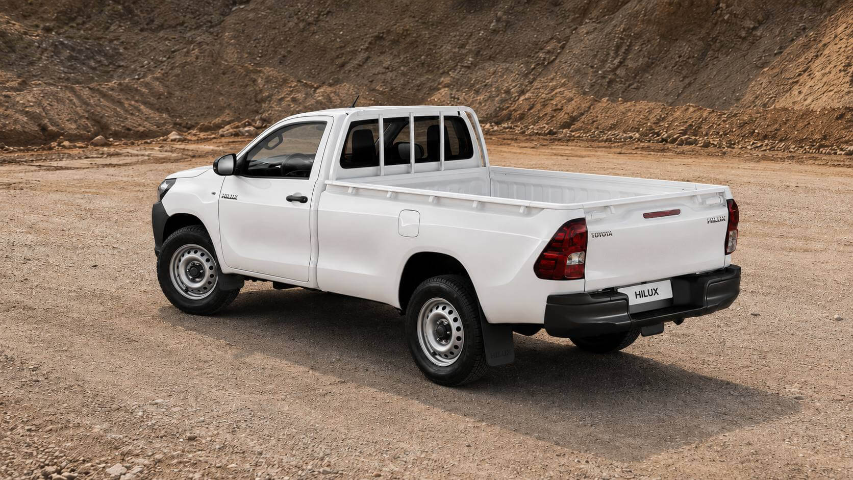 Toyota Hilux 2.8 turbodiesel 4x4 cabine simples 2019 na cor branca, vista em tres quartos traseira, parada sobre um patio de terra batida e cascalho sob luz natural. A picape tem cabine curta de duas portas seguida da cacamba longa, protetor de cabine tubular atras do vidro traseiro, rodas de aco sem calotas, para-barros com o nome HILUX, para-choque traseiro escuro e o letreiro TOYOTA na tampa da cacamba; a placa traseira mostra apenas HILUX.
