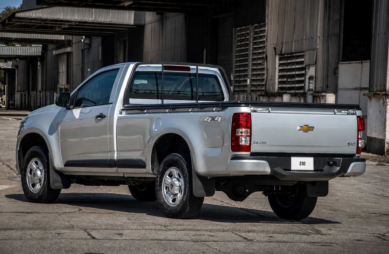 Chevrolet S10 LS 2.8 turbodiesel 4x4 cabine simples 2019 na cor prata, vista em tres quartos traseira, parada sobre o piso de concreto rachado de um patio industrial, com galpoes de estrutura metalica escura ao fundo. A picape tem duas portas e cacamba longa, rodas de aco prateadas, para-choque e retrovisores escuros, para-barros atras das rodas, santantonio baixo de tubo preto sobre a cabine e antena no teto. Na lateral da cacamba aparece o emblema 4x4 e, na tampa traseira, a gravata dourada da Chevrolet, a inscricao 2.8 CTDi e o logotipo S10; a placa traseira traz apenas a inscricao S10. A luz natural quente do fim da tarde ilumina a lateral do veiculo e projeta sua sombra no chao.