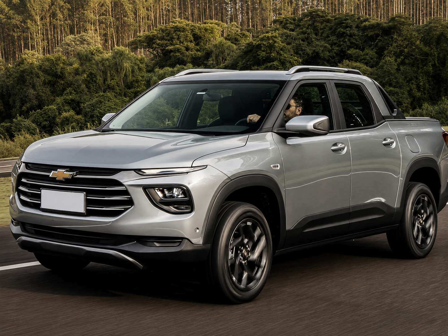 Chevrolet Montana LS 2023 na cor prata, picape de cabine dupla vista em três quartos dianteiro enquanto roda por uma estrada asfaltada, com o motorista ao volante e um paredão de mata verde ao fundo sob luz dourada de fim de tarde; a grade dianteira exibe o emblema gravata-borboleta da Chevrolet e a placa está em branco
