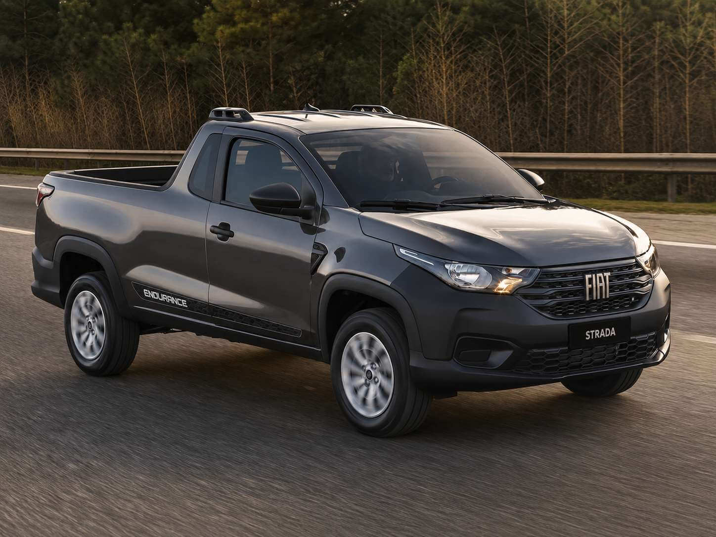 Fiat Strada Endurance Cabine Simples 2023 na cor cinza grafite, picape compacta de cabine simples vista em tres quartos dianteiro, rodando em uma estrada asfaltada com guard-rail e mata ao fundo sob luz dourada de fim de tarde; destaque para a cacamba grande com protetor, as rodas de aco de 15 polegadas, o adesivo ENDURANCE na lateral e o logotipo FIAT na grade