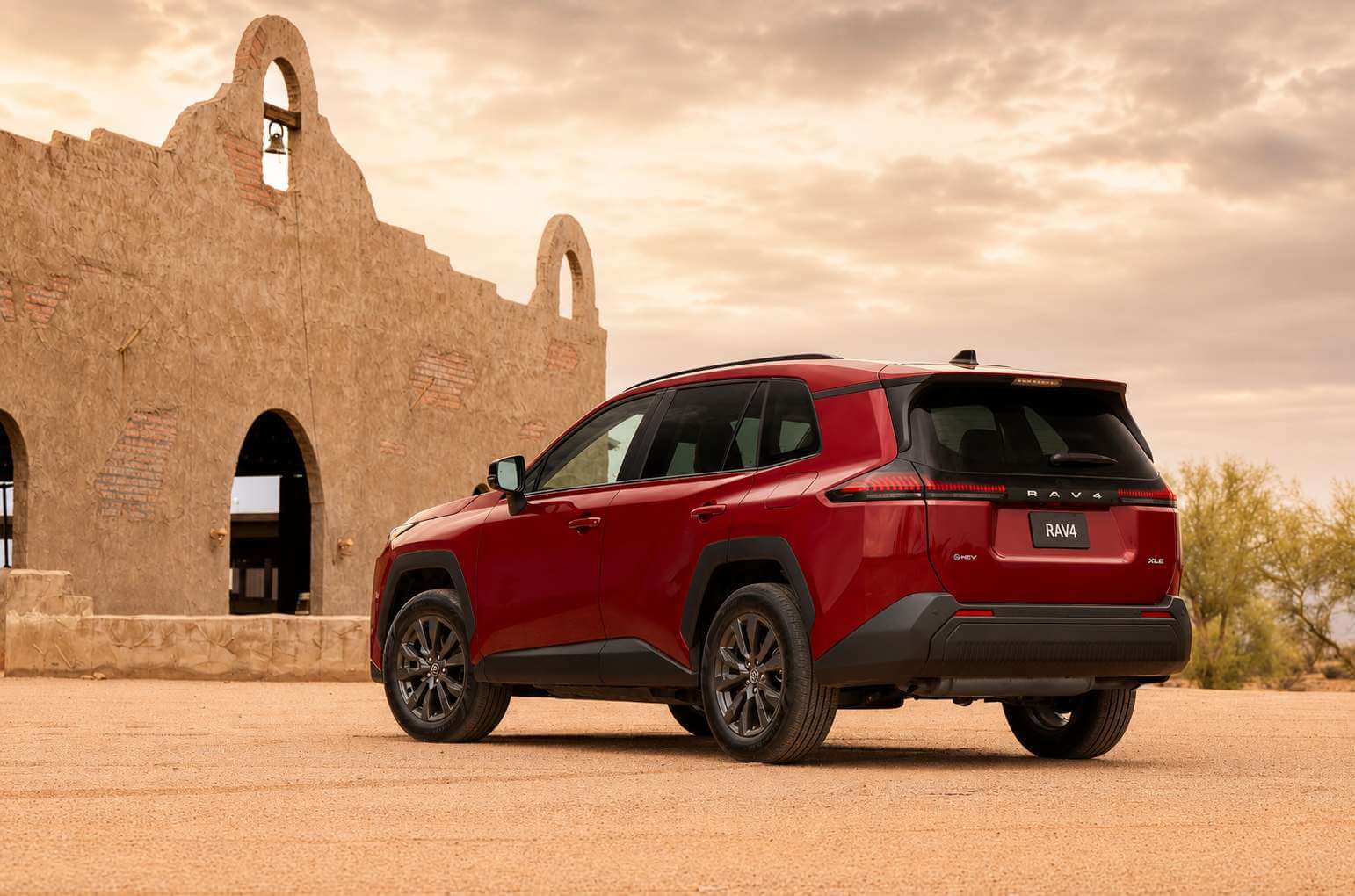 Toyota RAV4 híbrido na cor vermelha (Ruby Flare Pearl) visto em três quartos traseiro, parado num pátio de terra diante de uma antiga construção de adobe com arcos e um sino, sob céu nublado ao entardecer. Dá pra ver o lettering “RAV4” na tampa traseira, as rodas de liga escuras e os emblemas HEV e XLE.