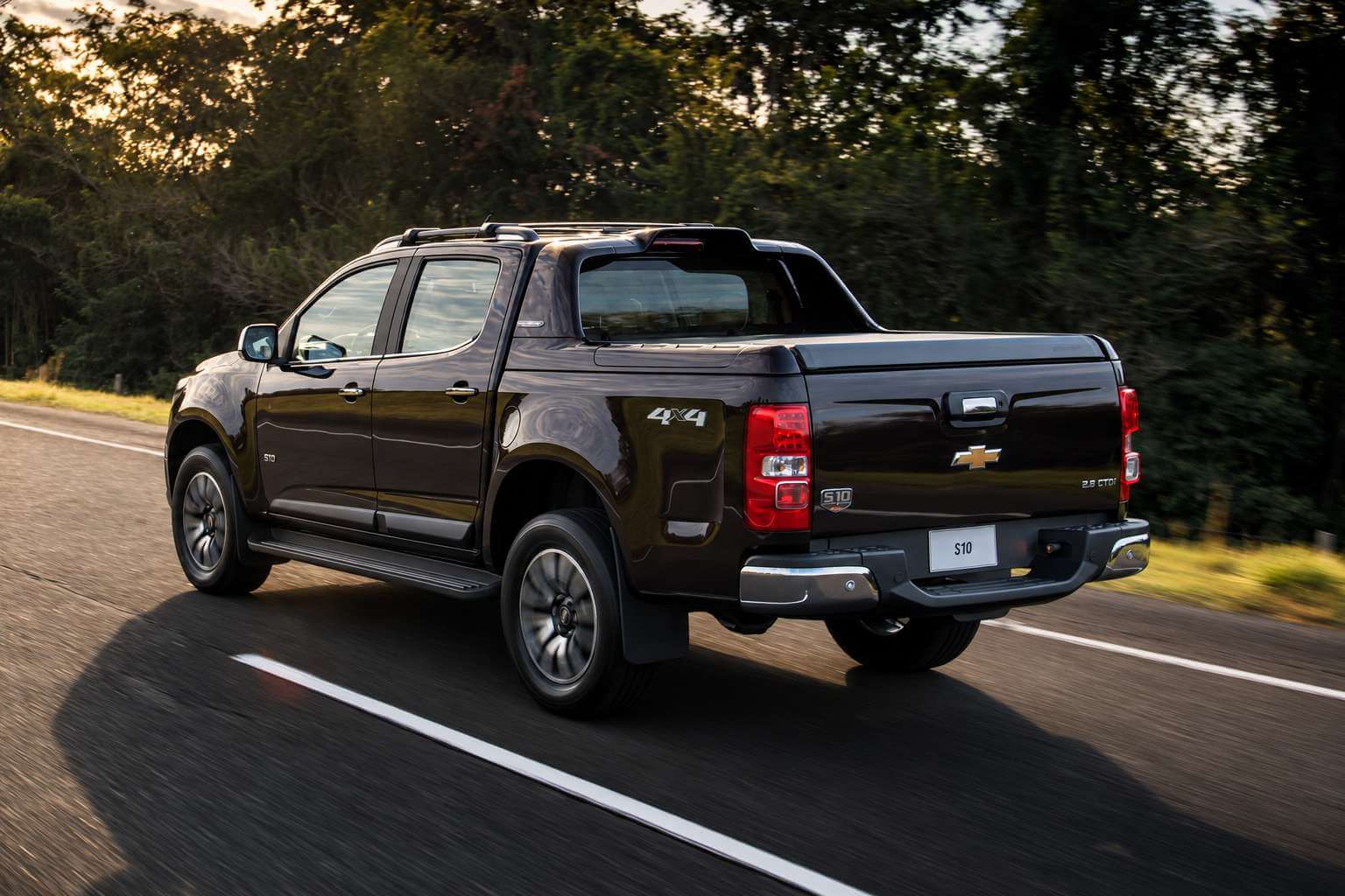 Chevrolet S10 LTZ 2.8 turbodiesel 4x4 cabine dupla 2018, na cor vinho escuro, vista em tres quartos traseiros enquanto roda por uma estrada asfaltada de pista simples ao entardecer; a luz baixa e dourada bate na lateral direita, o para-choque traseiro cromado, os estribos laterais, as barras de teto e as lanternas verticais aparecem em detalhe, e sao visiveis o emblema Chevrolet no meio da tampa da cacamba, a inscricao 2.8 CTDi a direita e o adesivo 4x4 na lateral da cacamba; ao fundo, mata verde desfocada pelo movimento