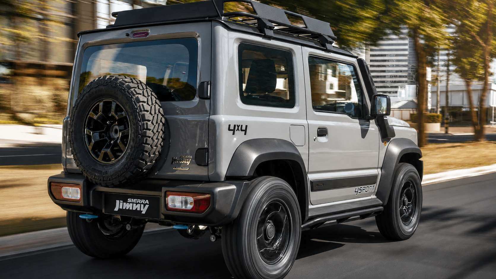 Suzuki Jimny Sierra 4Sport 2023 prata visto de tras em tres quartos, rodando por uma avenida arborizada com o fundo desfocado pela velocidade; o jipinho de tres portas traz estepe externo preso na tampa traseira, rack de teto preto, letreiro 4x4 na carroceria, faixa 4SPORT na lateral e pneus de uso misto, sob luz quente de fim de tarde