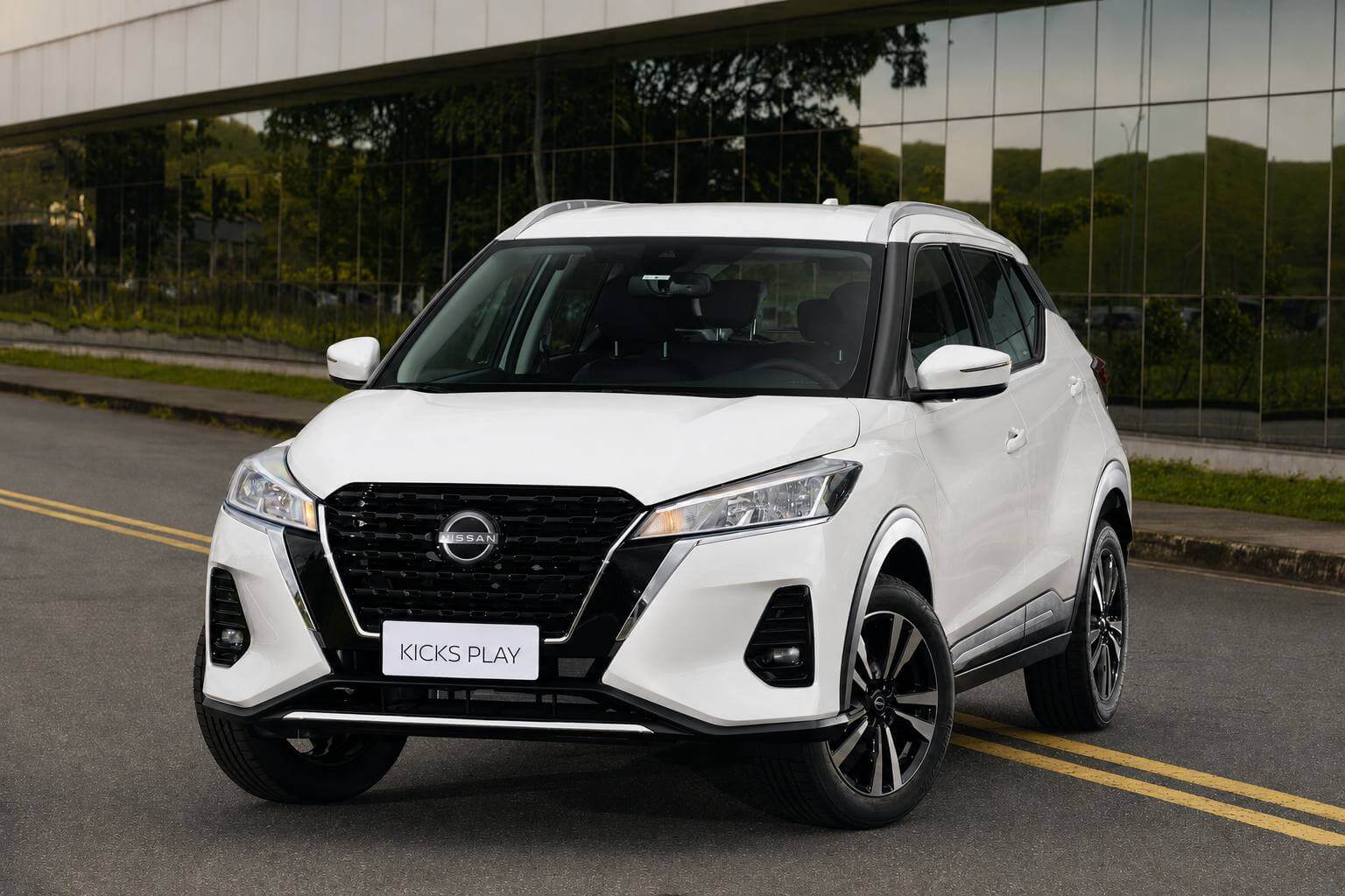 Nissan Kicks Play 2025 branco, visto de frente em tres quartos, parado em uma rua asfaltada com faixa amarela dupla ao lado de um predio de fachada espelhada e vegetacao verde ao fundo, em dia nublado; na placa dianteira le-se apenas KICKS PLAY