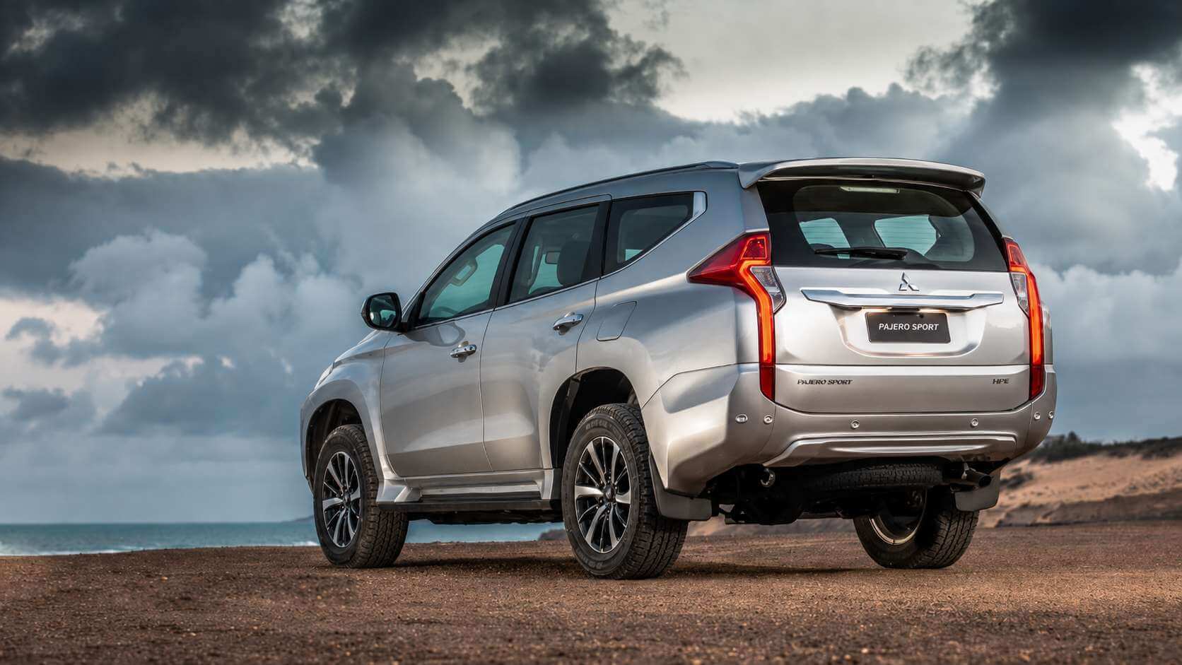 Mitsubishi Pajero Sport HPE prata fotografado em três quartos traseiro, parado sobre terreno de terra batida à beira-mar ao entardecer, com céu carregado de nuvens escuras ao fundo. Aparecem a tampa traseira com o emblema de três diamantes da Mitsubishi, o letreiro PAJERO SPORT e a insígnia HPE, as lanternas verticais em L, o aerofólio de teto, os estribos laterais e as rodas de liga diamantadas com pneus de uso misto.