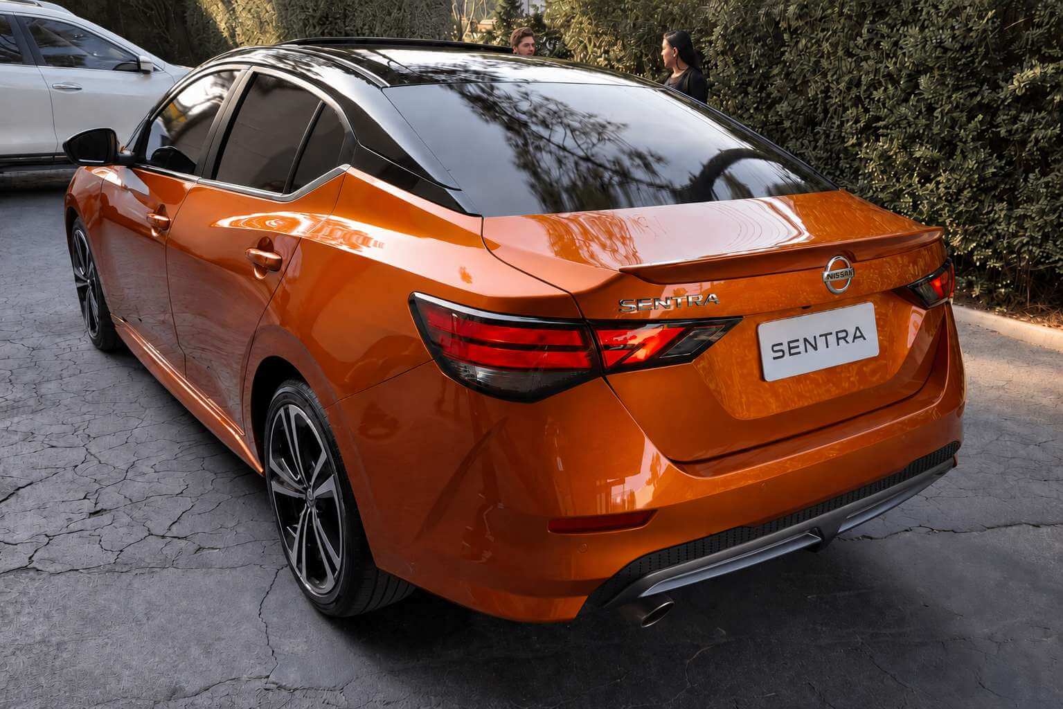 Nissan Sentra 2023 laranja metalico com teto preto, visto de tras em tres quartos, parado sobre um patio de asfalto rachado em dia de sol; aparecem as lanternas escurecidas acesas, o aerofolio discreto na tampa do porta-malas, o emblema da Nissan e o letreiro SENTRA na tampa, e rodas de liga escuras de raios duplos; ao fundo ha uma cerca viva alta, arvores e duas pessoas conversando; na placa traseira le-se apenas SENTRA