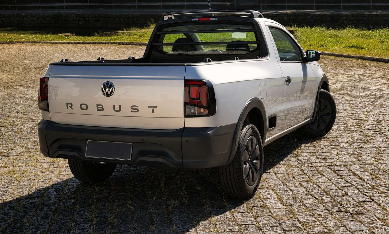 Volkswagen Saveiro Robust cabine simples prata vista em tres quartos traseiros, com o nome ROBUST em letras espacadas na tampa da cacamba, emblema da Volkswagen ao centro, santantonio preto sobre a cacamba, rodas escuras e parachoque traseiro preto, estacionada sobre piso de pedra portuguesa em dia de sol