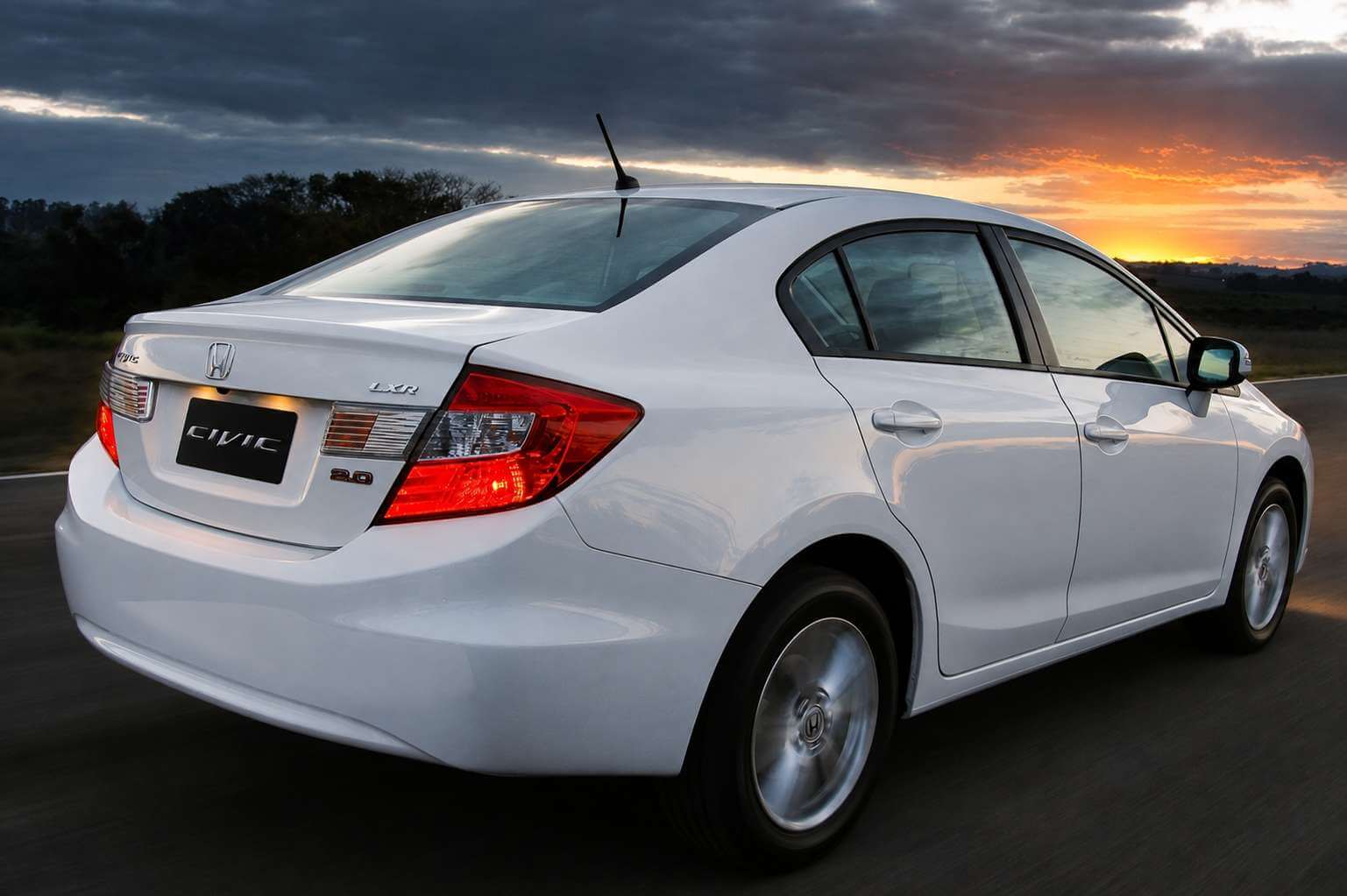 Honda Civic LXR 2.0 2014 branco, da geração G9, fotografado em três quartos traseiro enquanto roda por uma estrada de asfalto no fim da tarde. A traseira mostra as lanternas vermelhas alongadas em formato de bumerangue, o emblema da Honda, as inscrições LXR e 2.0 na tampa do porta-malas e a placa escrita CIVIC. Ao fundo, vegetação baixa na beira da pista e um céu de pôr do sol alaranjado com nuvens escuras.
