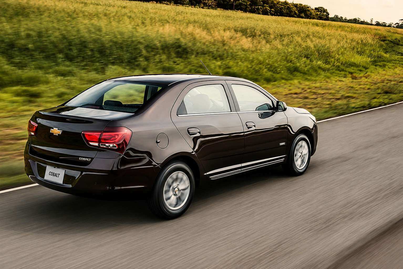 Chevrolet Cobalt 1.8 LTZ 2016 na cor marrom escuro metalico, visto de tras em tres quartos, rodando por uma estrada de pista simples em fim de tarde, com um barranco de mato alto dourado pela luz do sol ao fundo. O asfalto e a vegetacao aparecem borrados pelo movimento. Da para ver as lanternas vermelhas verticais, o logo dourado da Chevrolet e o nome Cobalt em letras cromadas na tampa do porta-malas, as rodas de liga de cinco raios, a faixa cromada na lateral e a placa escrita COBALT.
