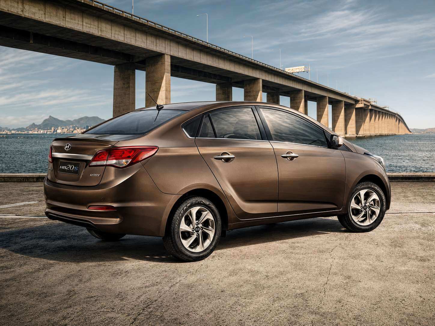 Hyundai HB20S 1.6 automático 2016 na cor marrom bronze metálico, visto de trás em três quartos, parado sobre um piso de concreto à beira-mar; ao fundo, uma longa ponte de vigas de concreto atravessa o mar em direção a morros e à cidade no horizonte, sob céu azul com nuvens finas. Dá para ver a lanterna traseira alongada, o emblema da Hyundai e o letreiro HB20S na tampa do porta-malas, além das rodas de liga leve de raios duplos