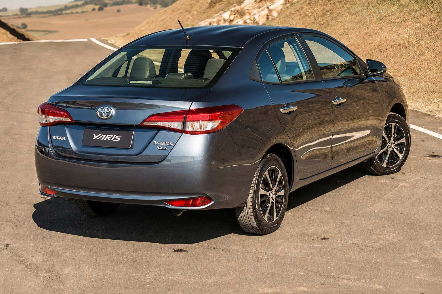 Toyota Yaris Sedan cinza-azulado visto em três quartos traseiros, parado em uma pista de asfalto com barranco de vegetação seca ao fundo sob luz natural forte; lanternas vermelhas acesas na tampa do porta-malas, emblema da Toyota e a inscrição Yaris no centro da traseira, rodas de liga leve escuras e ponteira de escape aparente.