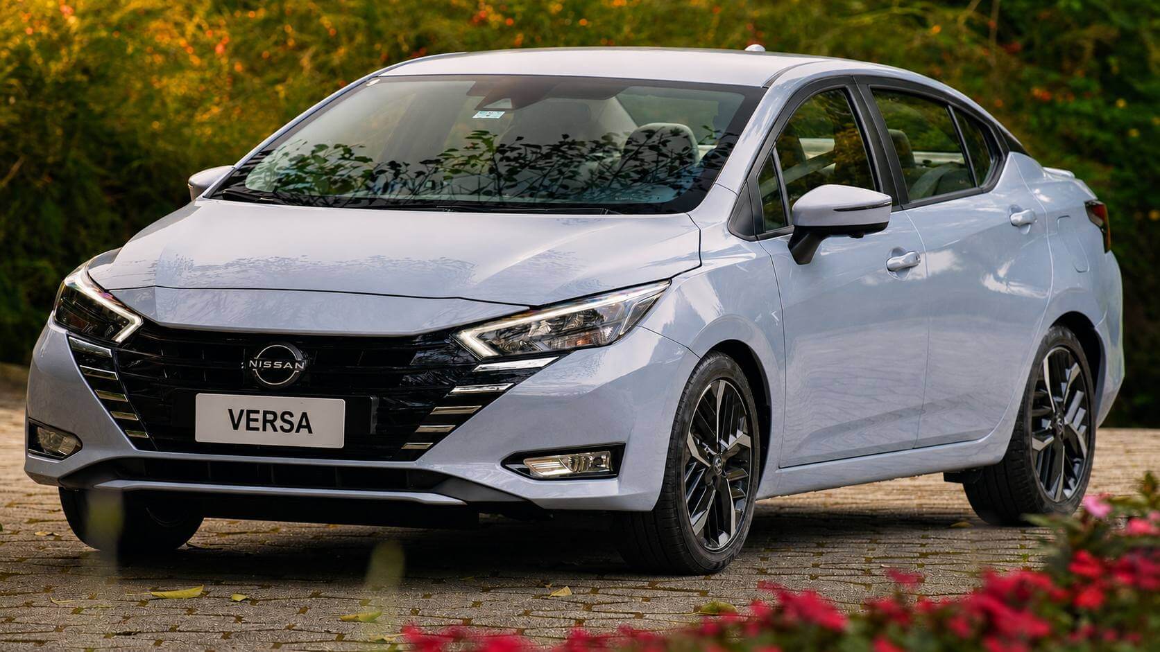 Nissan Versa 2023 cinza-prata visto em três quartos dianteiro, parado sobre piso de pedra portuguesa em meio a vegetação verde e flores rosa desfocadas em primeiro plano, sob luz natural quente; a placa dianteira traz apenas o nome VERSA.