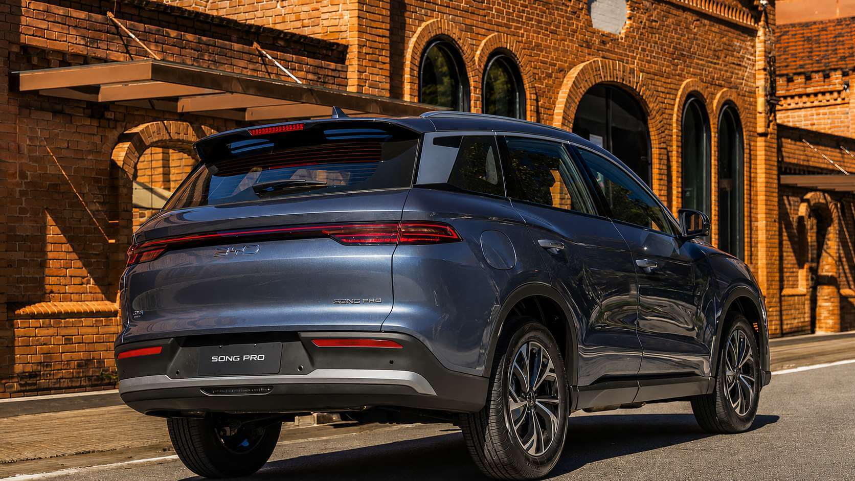 Traseira em três quartos de um SUV BYD Song Pro cinza-azulado parado numa rua de asfalto diante de um galpão antigo de tijolos aparentes com janelas em arco, sob luz de fim de tarde. A tampa traseira traz a faixa de lanterna vermelha de ponta a ponta com o letreiro BYD no centro, o letreiro SONG PRO em letras cromadas do lado direito e um pequeno emblema da versão no canto esquerdo; a placa preta traz apenas o nome do modelo, SONG PRO.