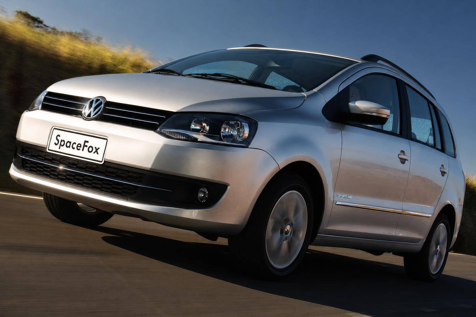 Volkswagen SpaceFox Sportline prata da fase pos-reestilizacao, linha 2011 a 2014, fotografada em movimento de tres quartos dianteiros e de angulo baixo em uma estrada asfaltada, com ceu azul limpo e vegetacao desfocada na lateral sob luz de fim de tarde. A perua traz a grade dianteira com filetes cromados e o emblema da Volkswagen no centro, farois com mascara escurecida, para-choque na cor da carroceria com farois de neblina redondos, retrovisores com setas integradas, friso cromado na lateral, a inscricao SPORTLINE na porta dianteira, longarinas no teto, rodas de liga leve aro 15 e a placa dianteira trazendo apenas o nome SpaceFox.