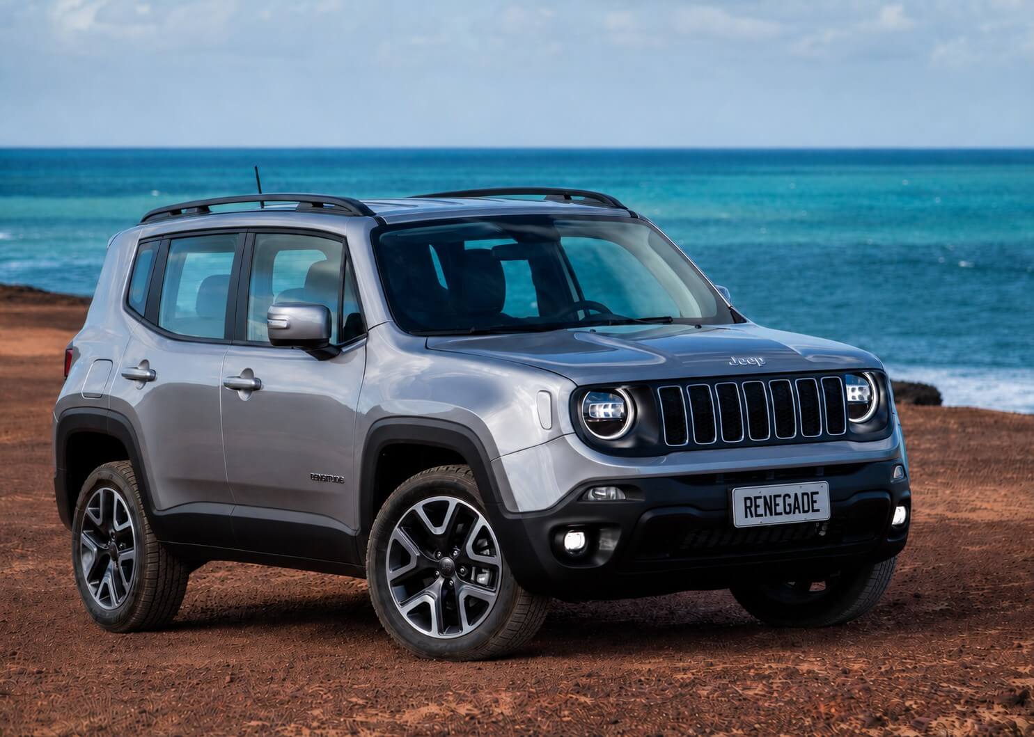 Jeep Renegade prata, SUV compacto a diesel, visto de tres quartos dianteiro num terreno de terra vermelha diante do mar, com grade de sete ranhuras, farois redondos e a placa escrita RENEGADE.