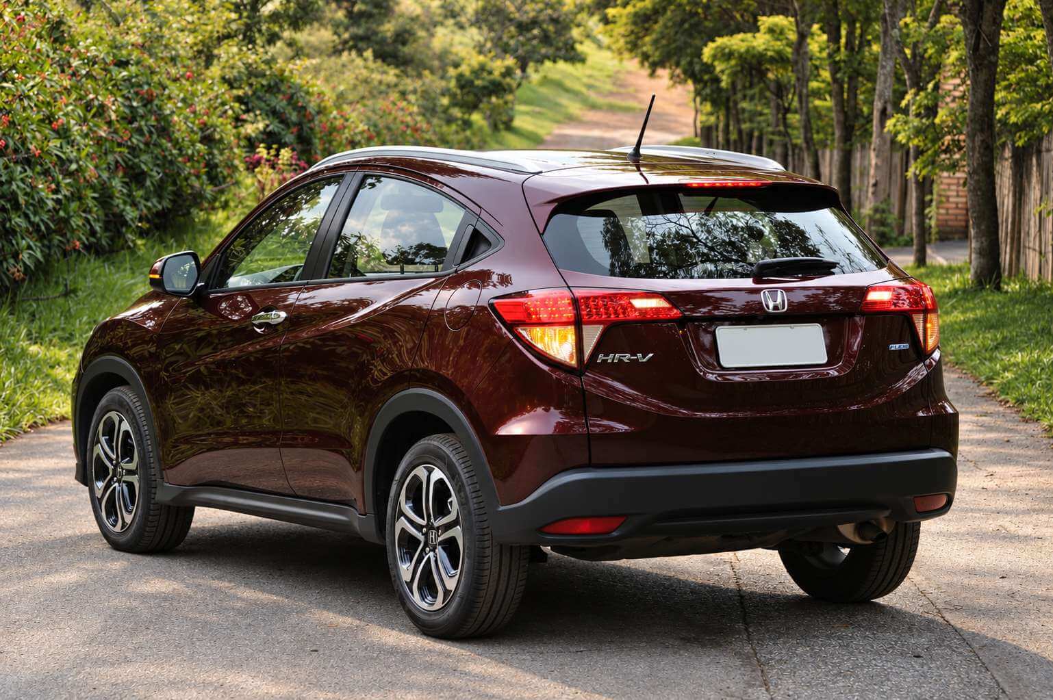 Honda HR-V EX 1.8 2016 na cor vinho metalico, fotografado de tres quartos traseiro, parado numa estrada de concreto cercada por arvores e vegetacao verde sob luz natural; aparecem as lanternas verticais em L, o emblema H da Honda e a letra HR-V na tampa do porta-malas, as rodas de liga de 17 polegadas e as barras longitudinais no teto.